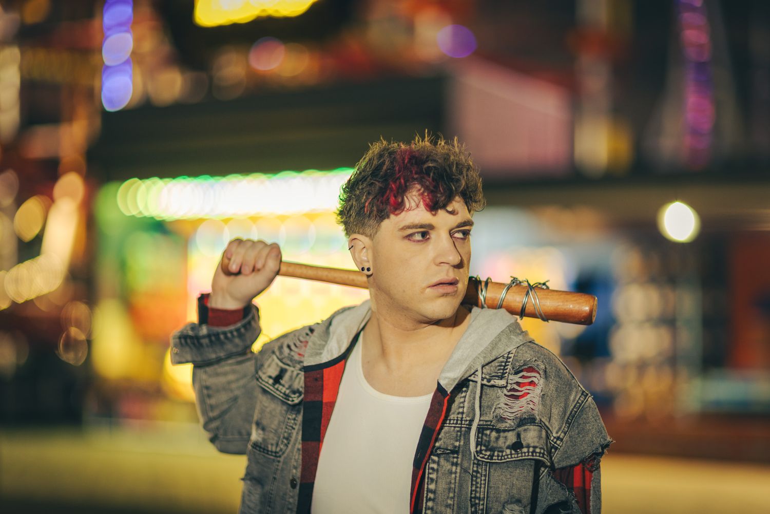 Young man with dyed curly hair rests a wooden baseball bat on his shoulder, wearing a denim jacket against glowing neon city lights.