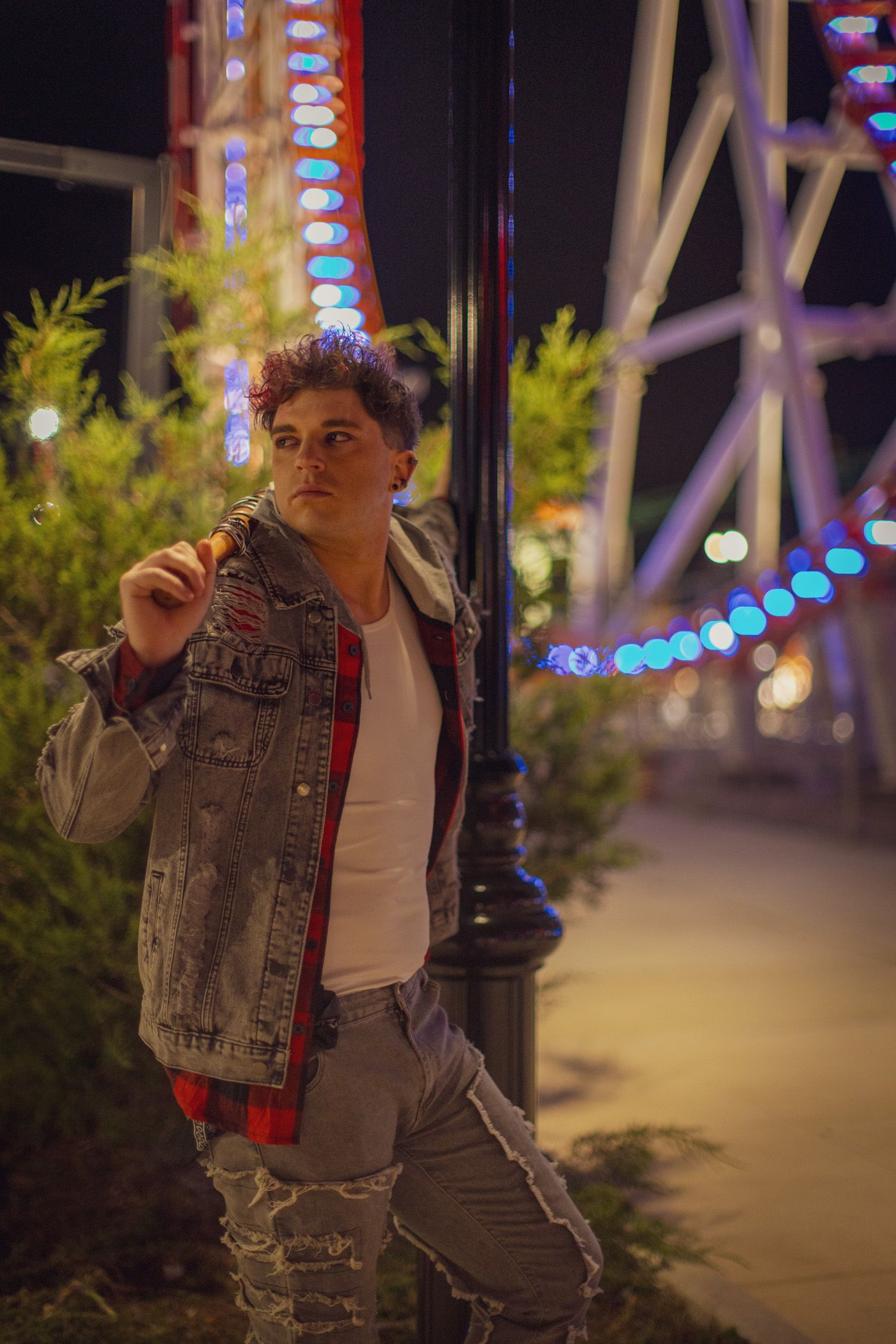Young man in a denim jacket, plaid shirt and ripped jeans leans on a lamp post at night near a lit amusement park ride.