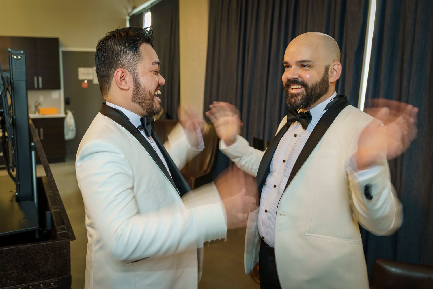 Two grooms in white tuxedos with black lapels laugh and clap together during wedding preparations.