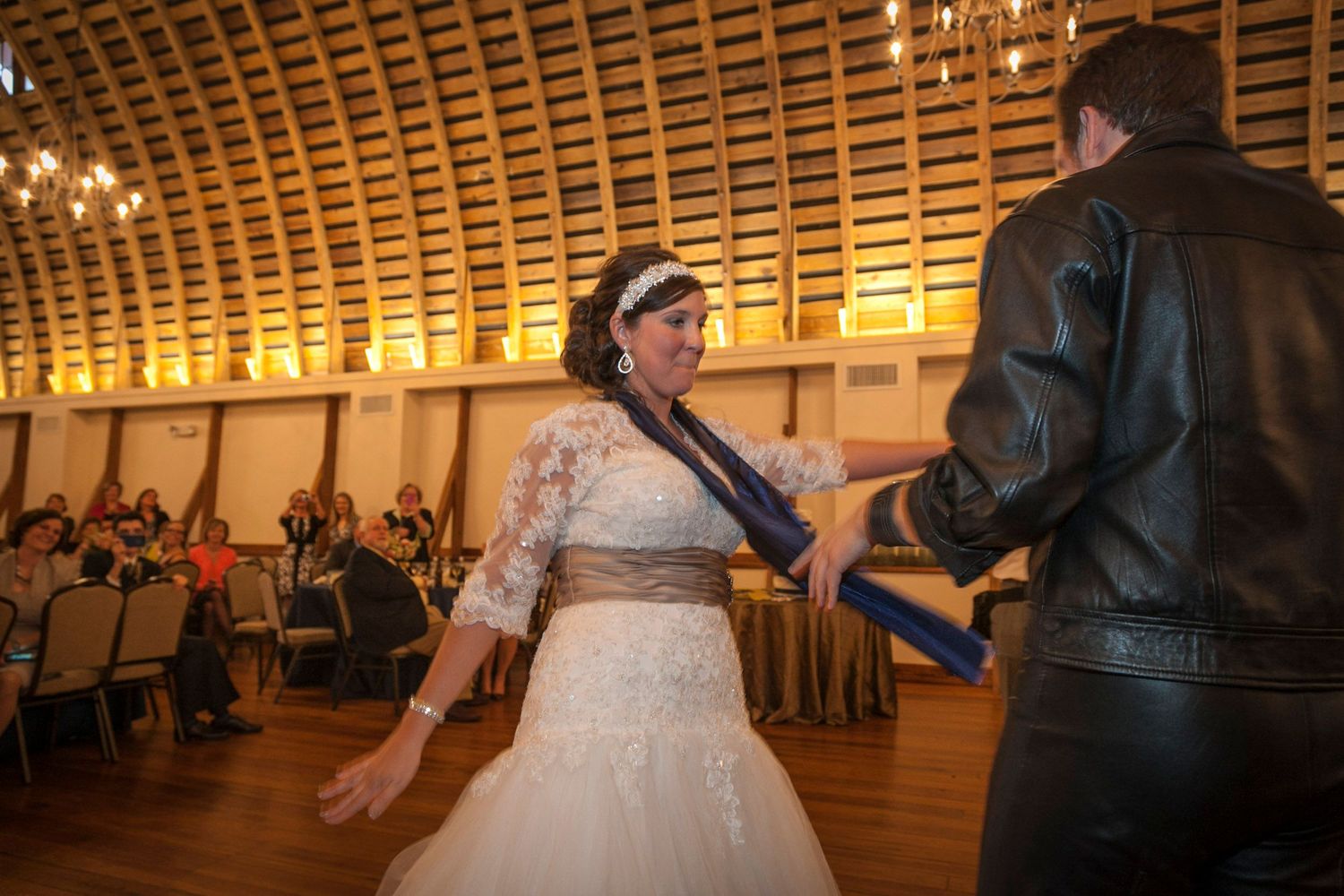 Bride in white ball gown dances with guest in leather jacket at elegant wedding reception.