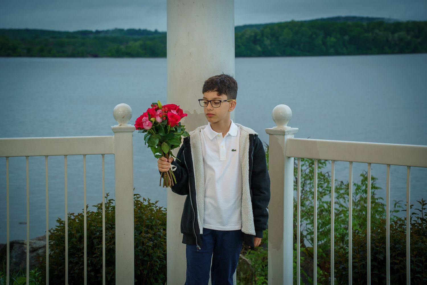 Young boy holding red roses bouquet standing on white railing balcony overlooking lake and green trees.