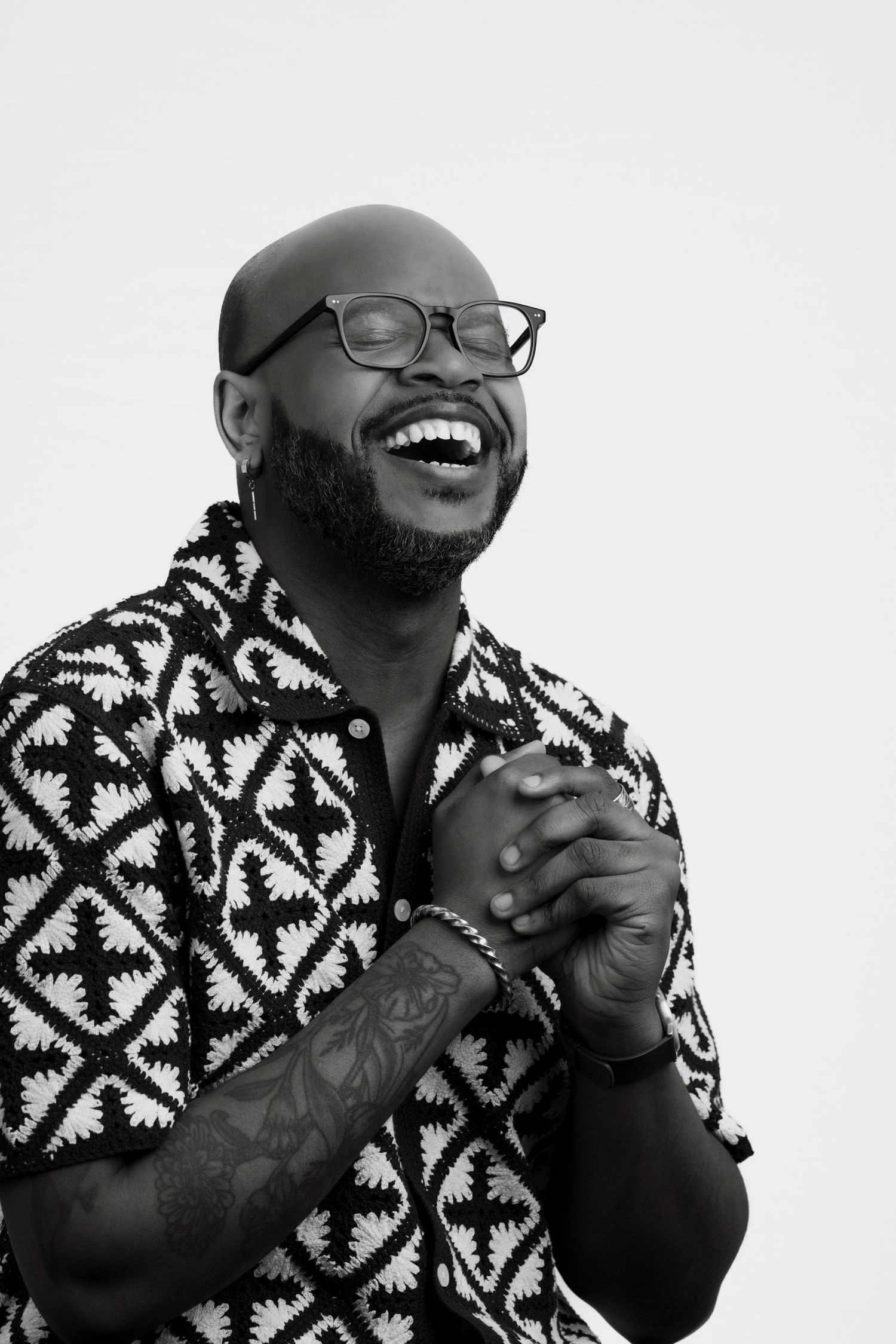 Black and white portrait of a bald man with glasses and beard laughing joyfully, wearing patterned shirt with tattoos.