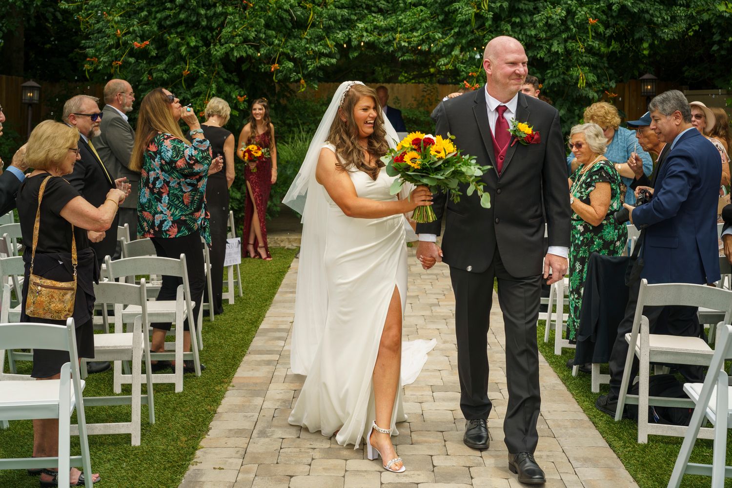 Newlywed couple walks down outdoor aisle after wedding ceremony, bride in white gown holding colorful bouquet.