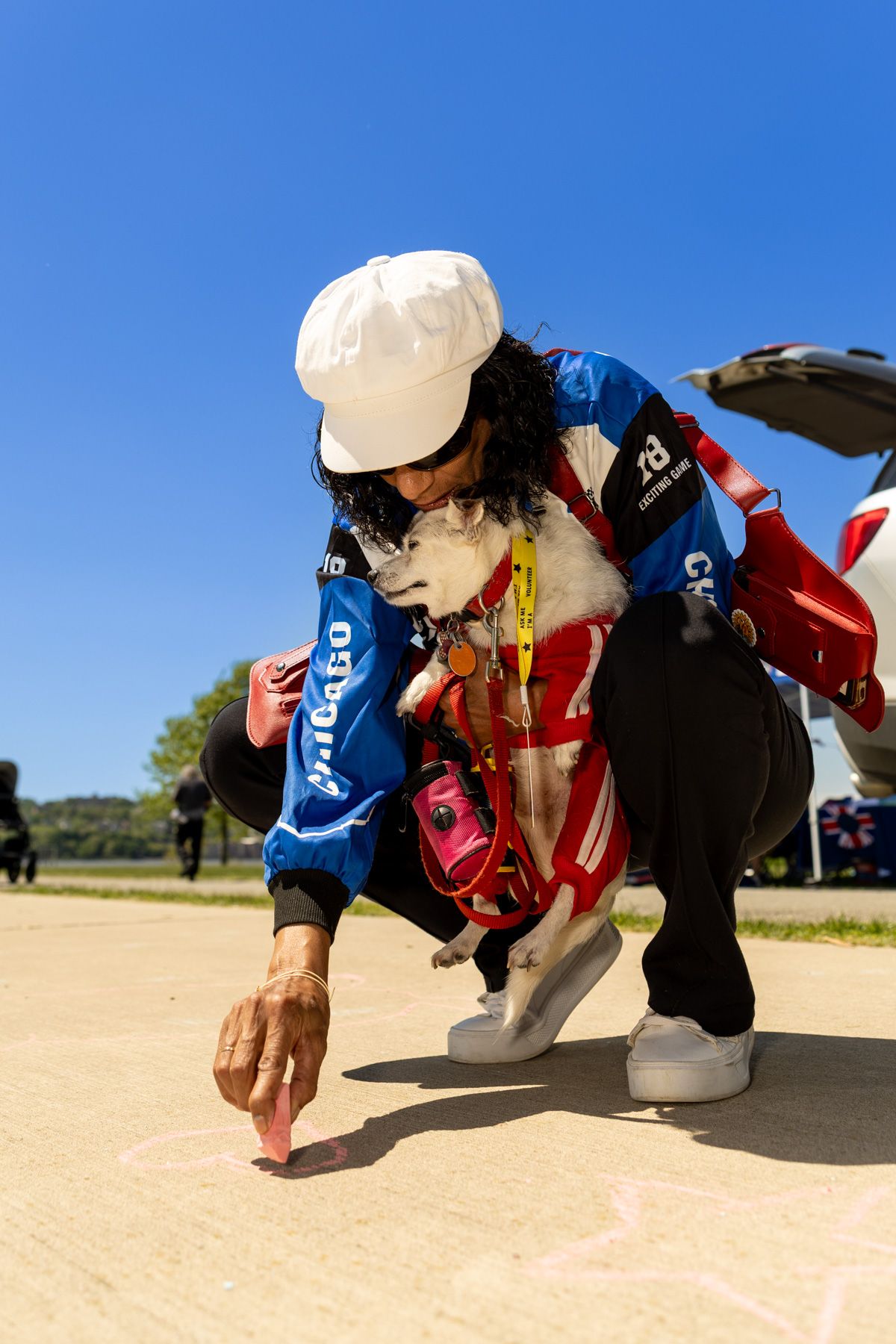 Woman in blue jacket and white cap crouches with small dog in red harness on pavement under clear blue sky.