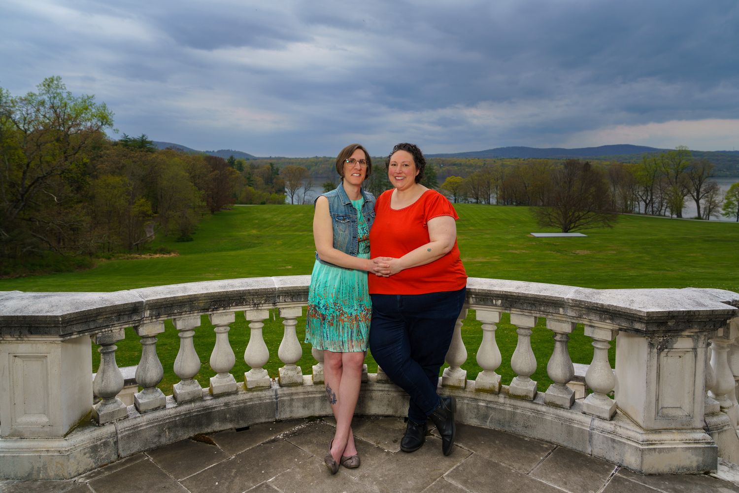 Two women pose on a stone balcony overlooking a lush green valley under dramatic cloudy skies.