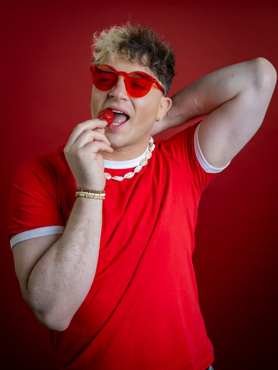 Man in red t-shirt and red heart sunglasses eating a strawberry against a bold red background.