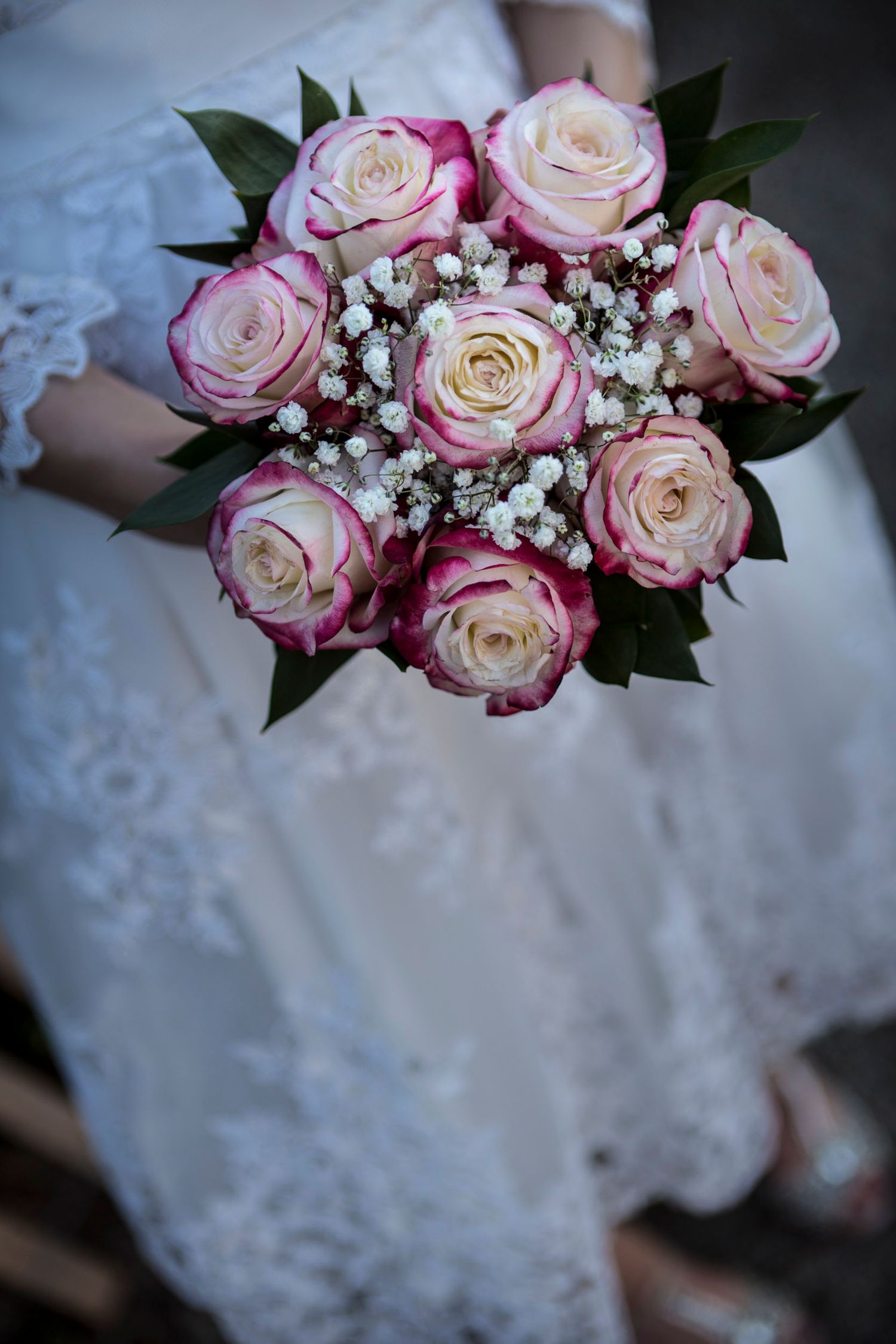 Bride in white lace dress holding a bridal bouquet of pink and cream roses with baby's breath.