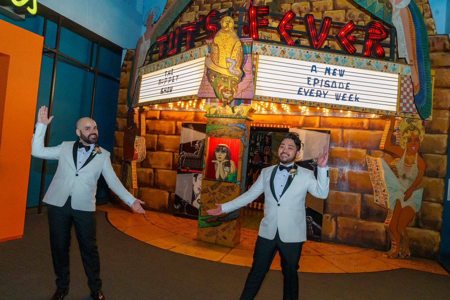 Two grooms in matching white tuxedos pose joyfully in front of a colorful theater marquee display.