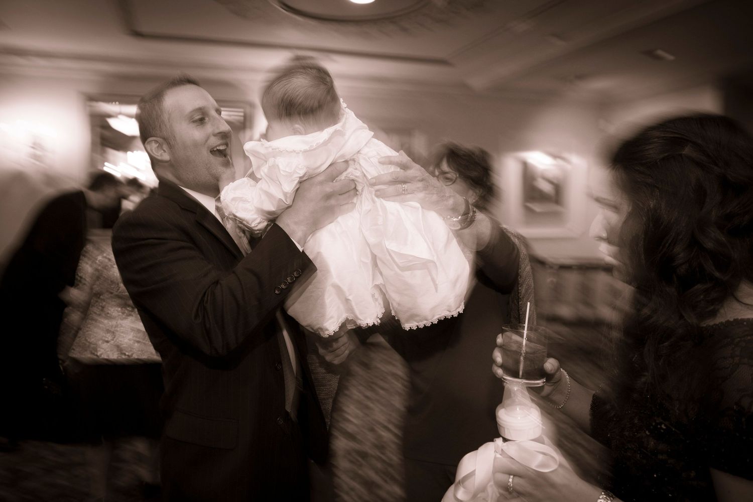 Man in formal attire joyfully lifting a baby in white christening gown at a reception.
