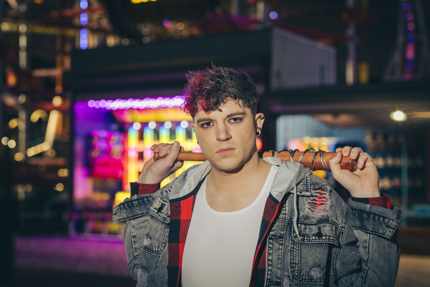 Young man with tousled dark hair wearing a denim jacket over a plaid shirt and white tank top poses against a neon-lit arcade background.