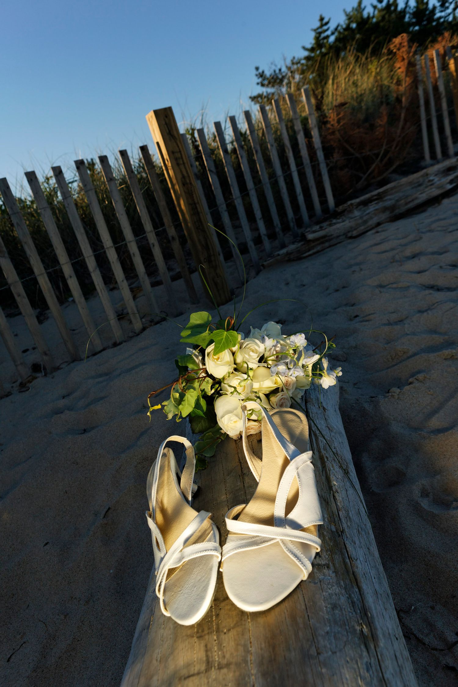 White bridal heels and flower bouquet resting on driftwood at beach sunset.