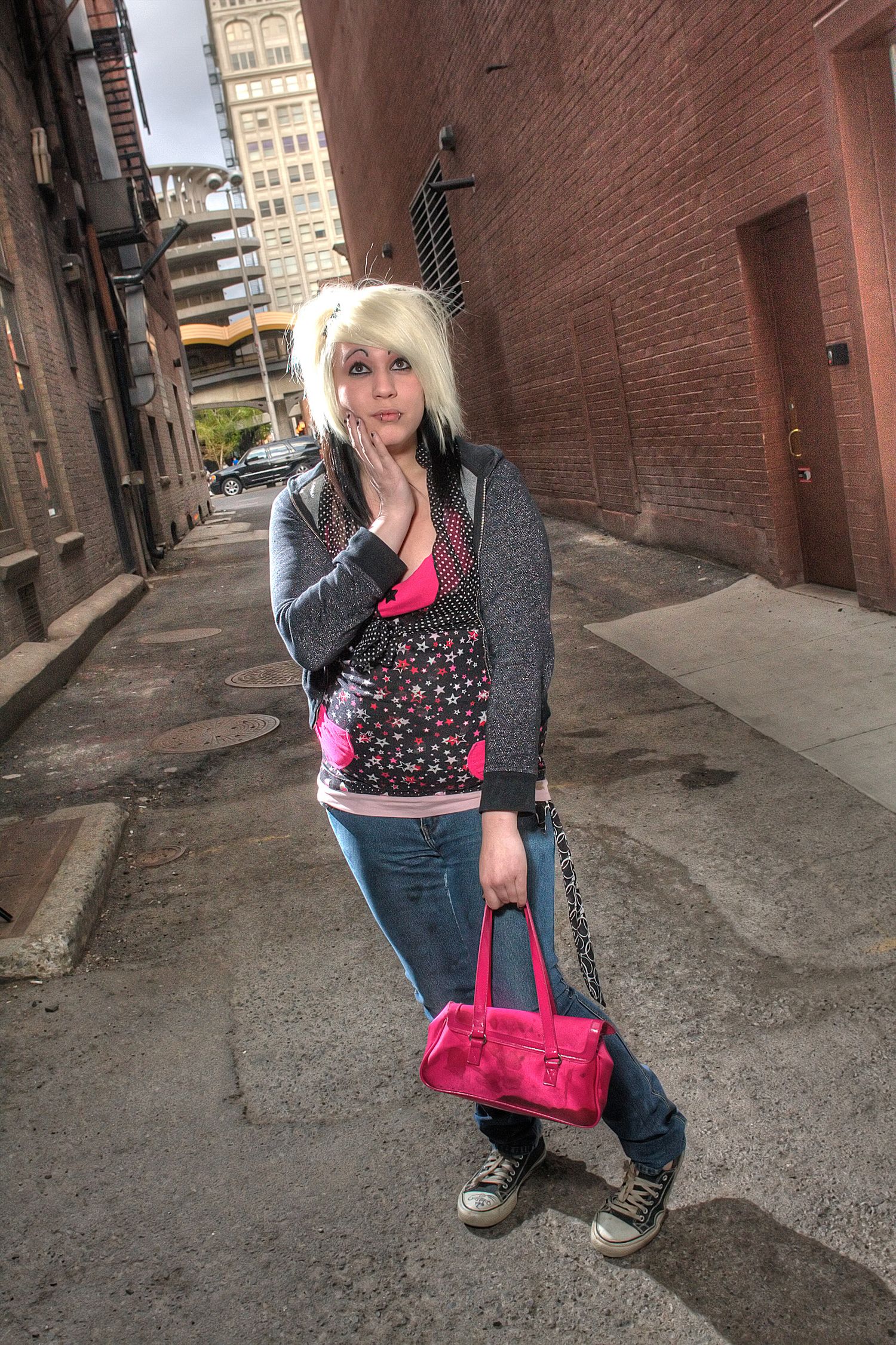 Blonde woman in pink and gray outfit holding pink purse in urban alley.