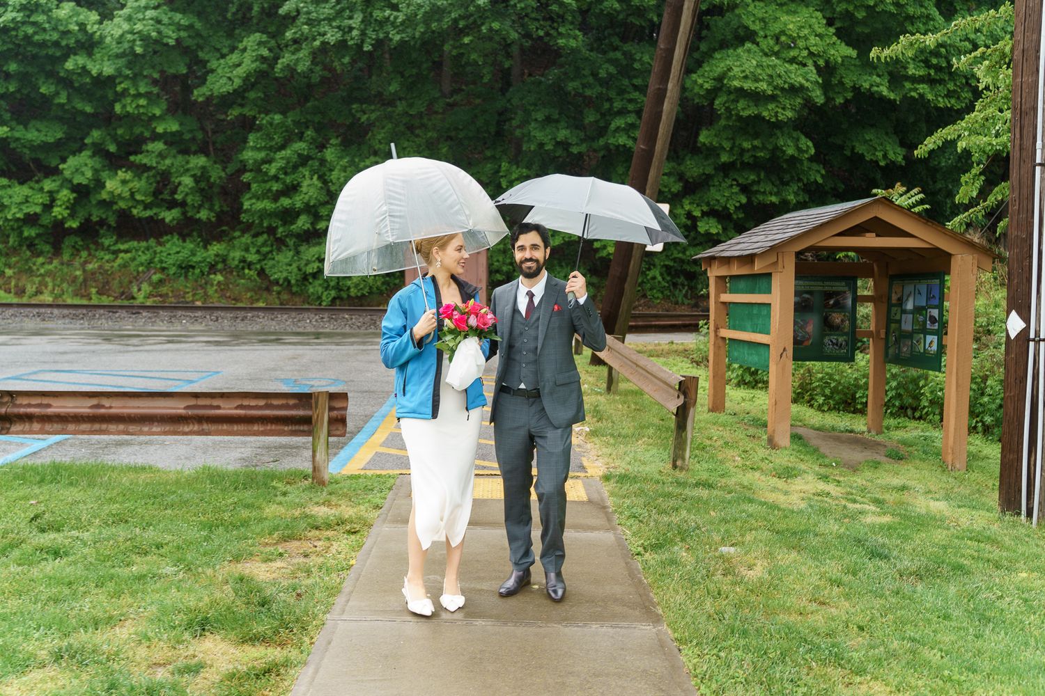 Couple walking under clear umbrellas on a rainy wedding day, bride holding red flowers, groom in gray suit, lush green background.