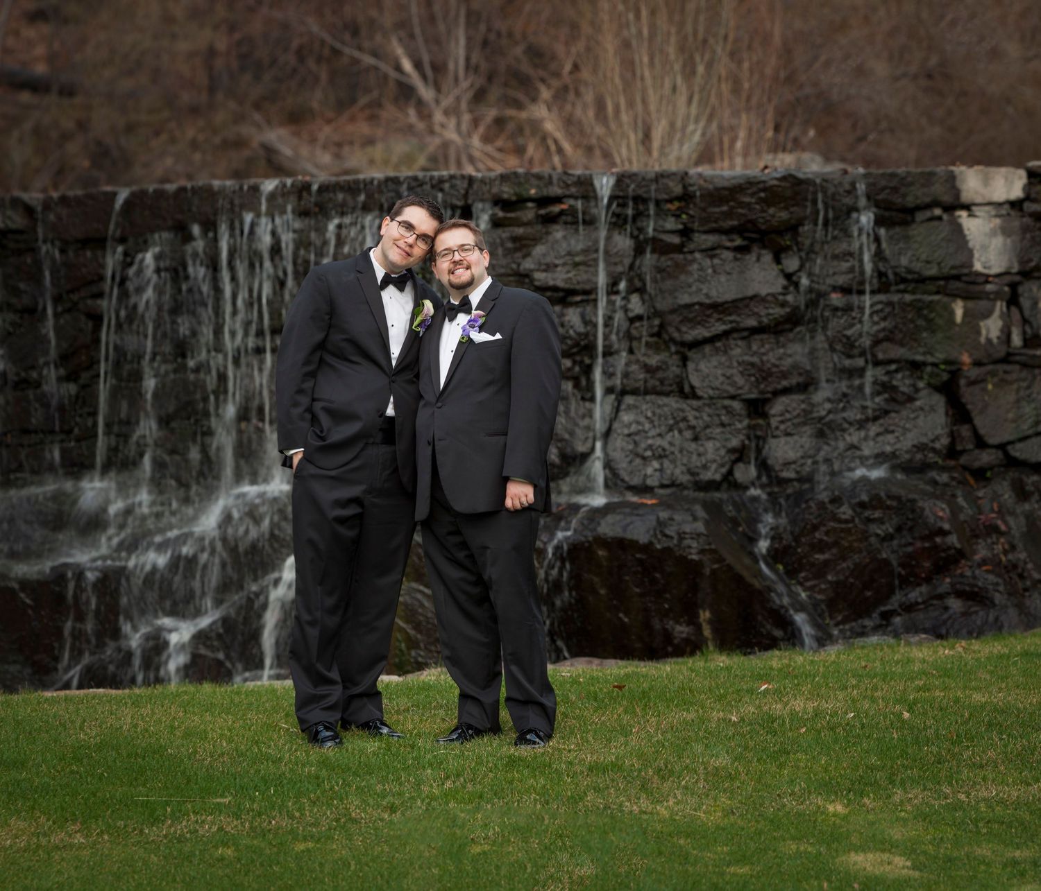 Two grooms in tuxedos pose together on green grass in front of a stone waterfall backdrop.