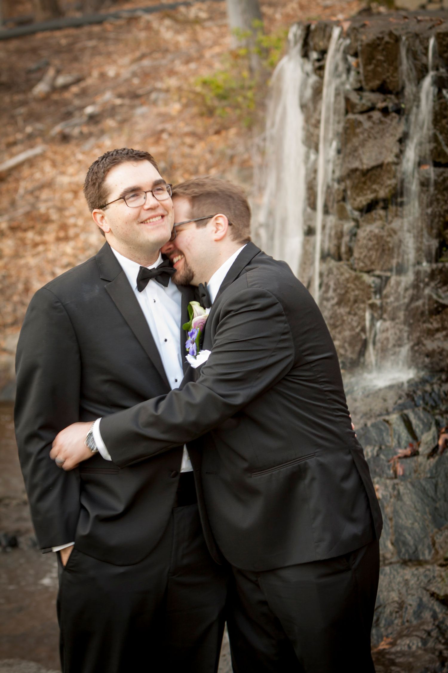 Two grooms in black tuxedos sharing a tender moment by a stone waterfall at their wedding.