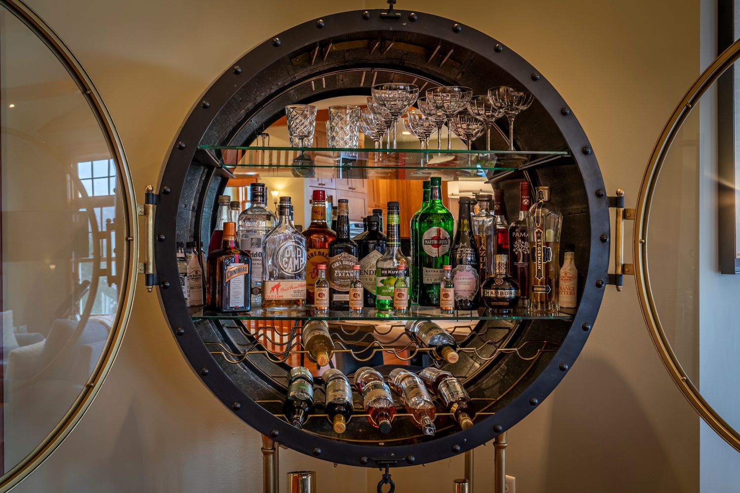 A circular porthole-style wall bar display filled with liquor bottles and glassware on wooden shelves.