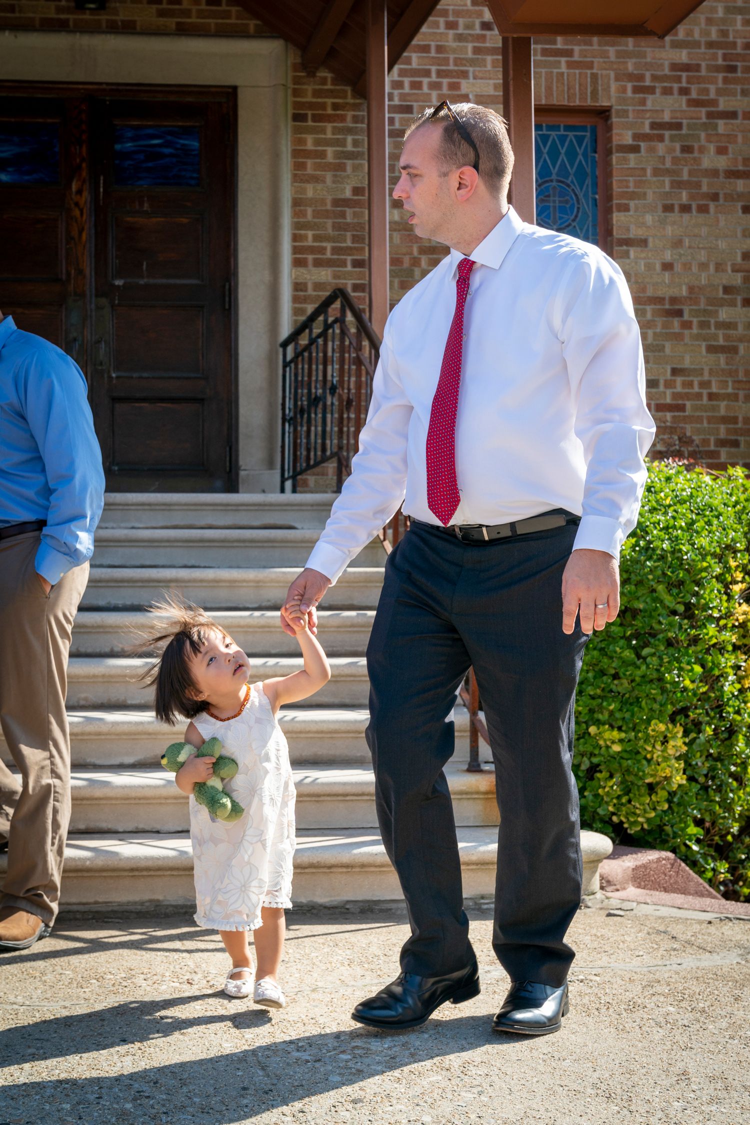 Man in white shirt and red tie holds hands with young girl in white dress outside brick church steps.