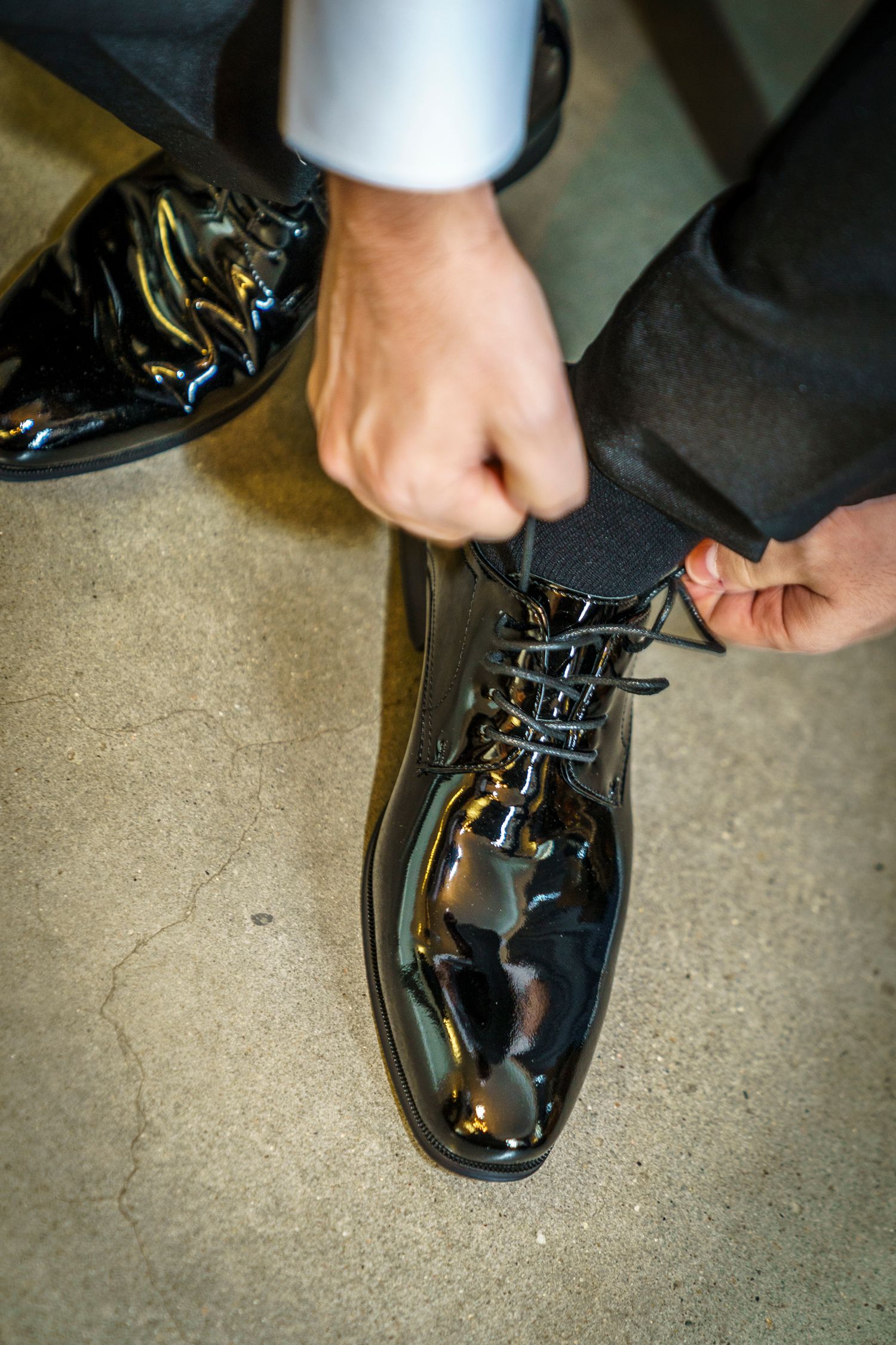 Man tying shiny black patent leather dress shoes while wearing a formal suit and white dress shirt.