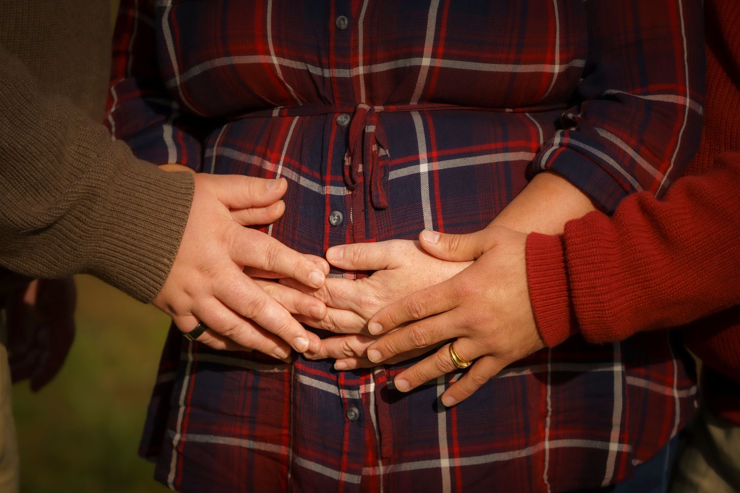 Couple tenderly holding hands on pregnant belly wearing plaid shirt, maternity photo outdoors.