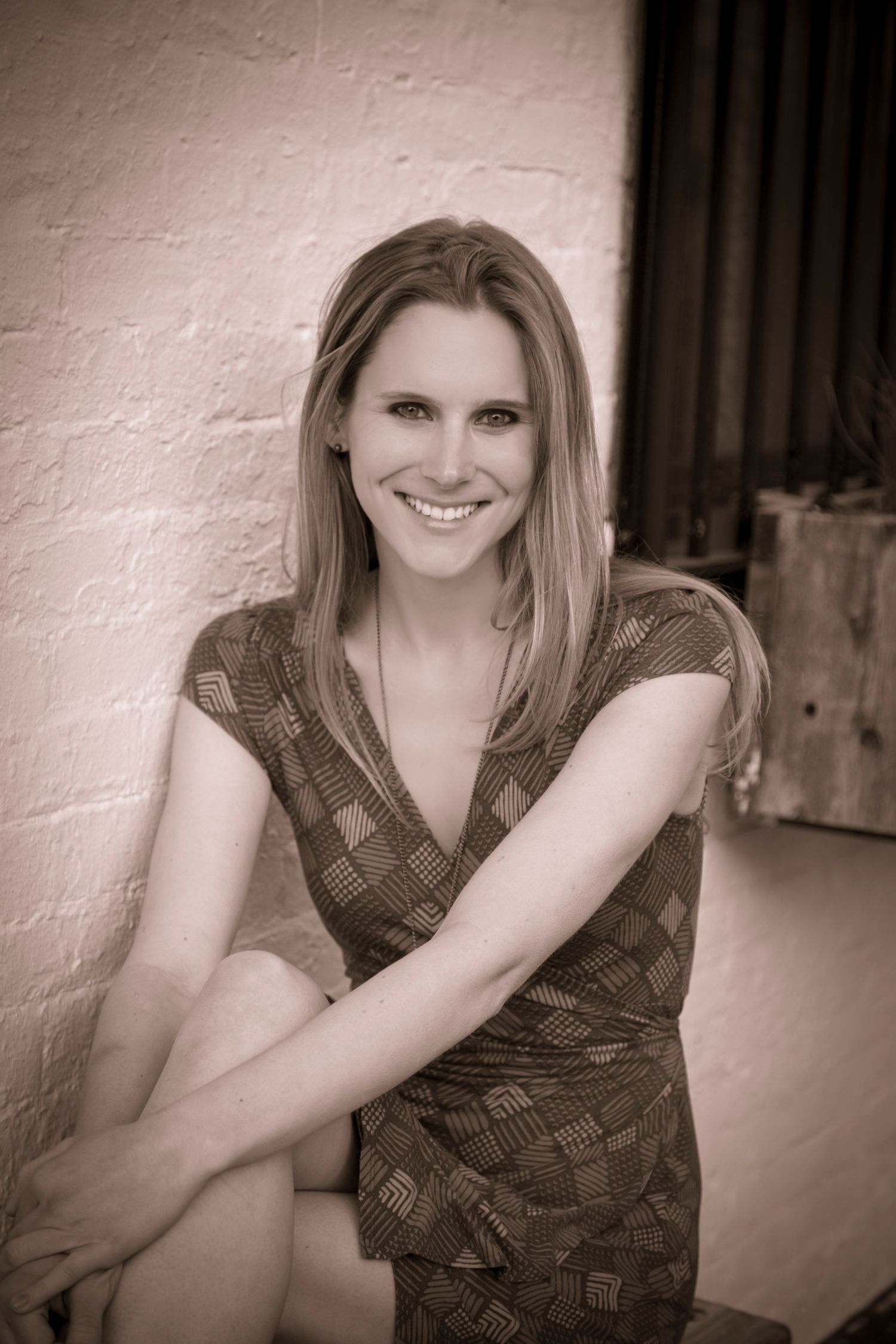 Smiling blonde woman in patterned dress sitting against a brick wall in a sepia-toned portrait photo.