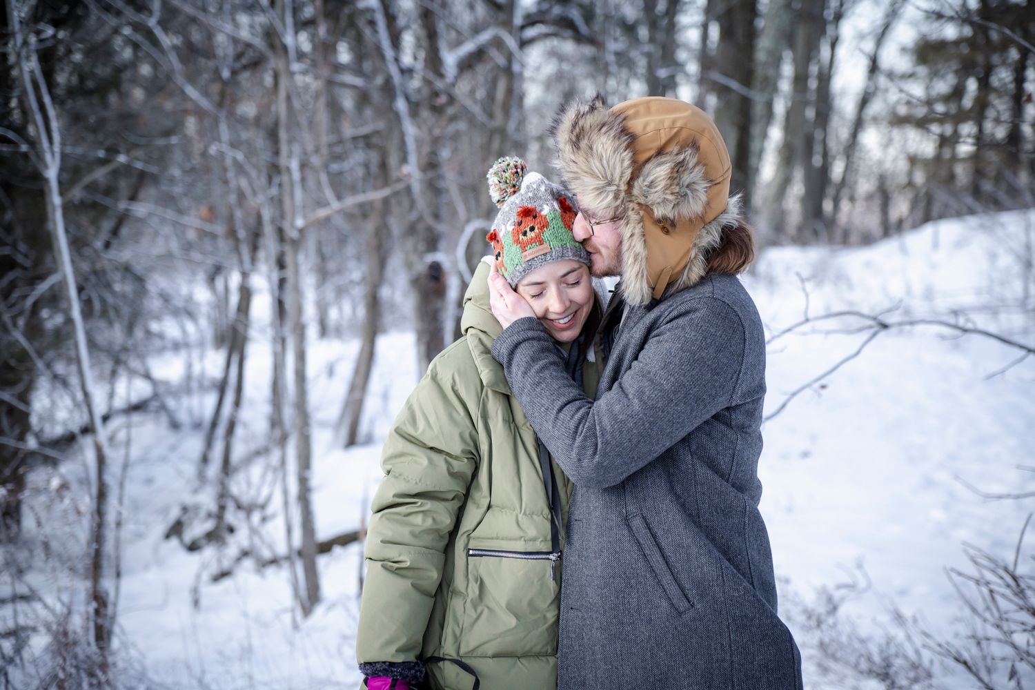 Two people in winter hats embrace warmly in a snowy forest, wearing cozy jackets on a cold winter day.