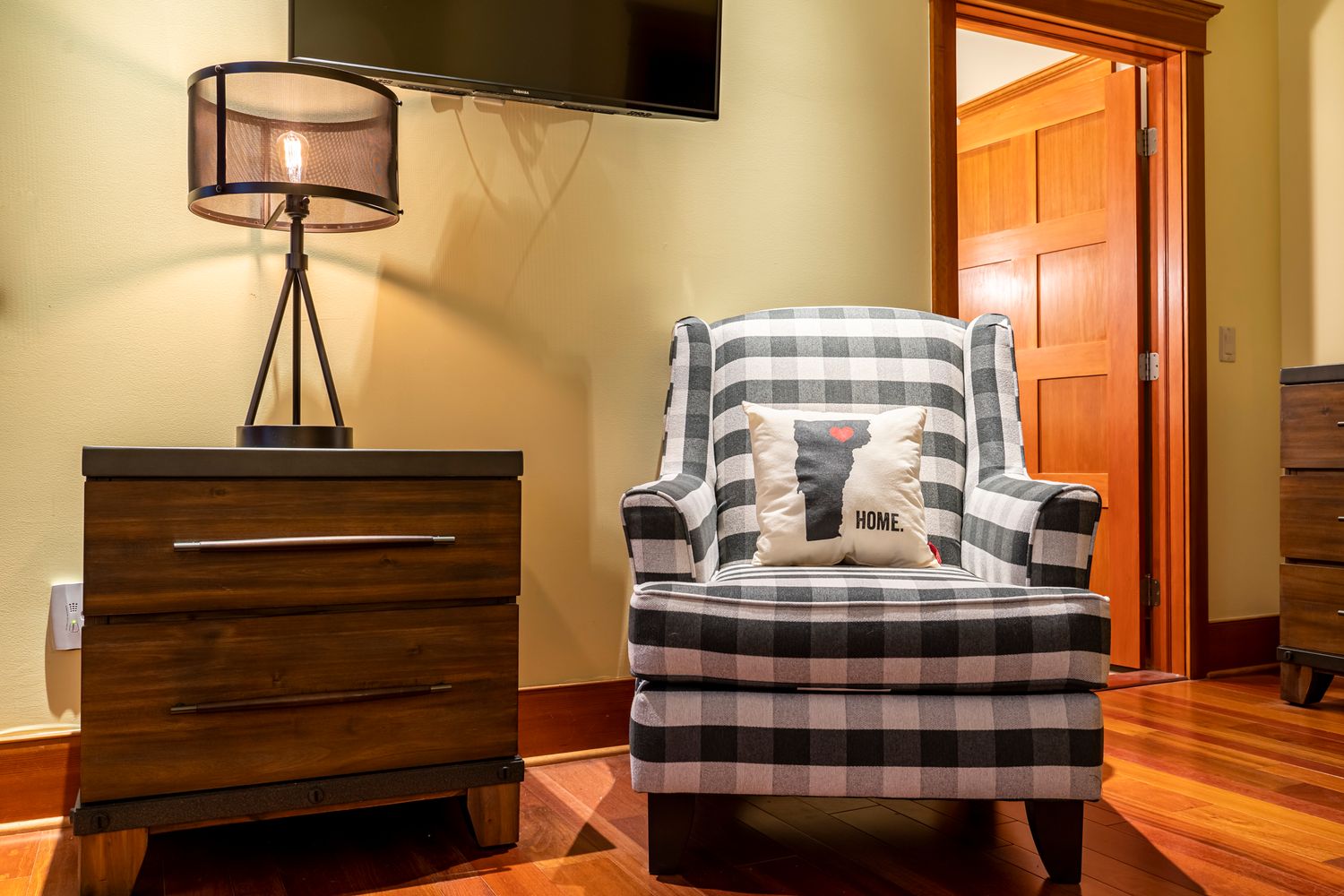 Cozy reading nook with a plaid armchair, wooden nightstand, and industrial lamp beside an open doorway.
