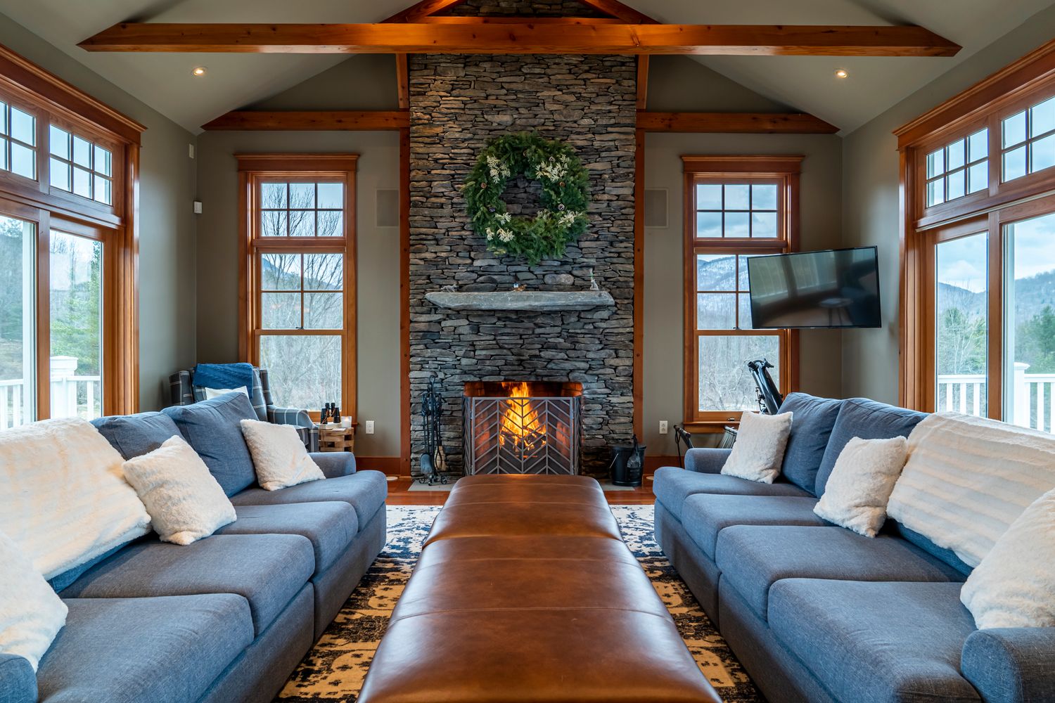 Cozy living room with stone fireplace, blue sofas, wood beams, and mountain views through large windows.