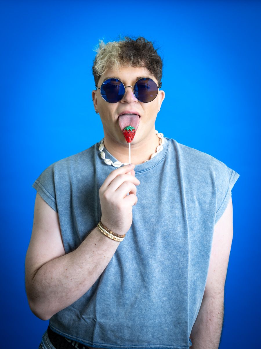 Young man with round sunglasses licking a strawberry lollipop, wearing a gray sleeveless shirt against a blue background.