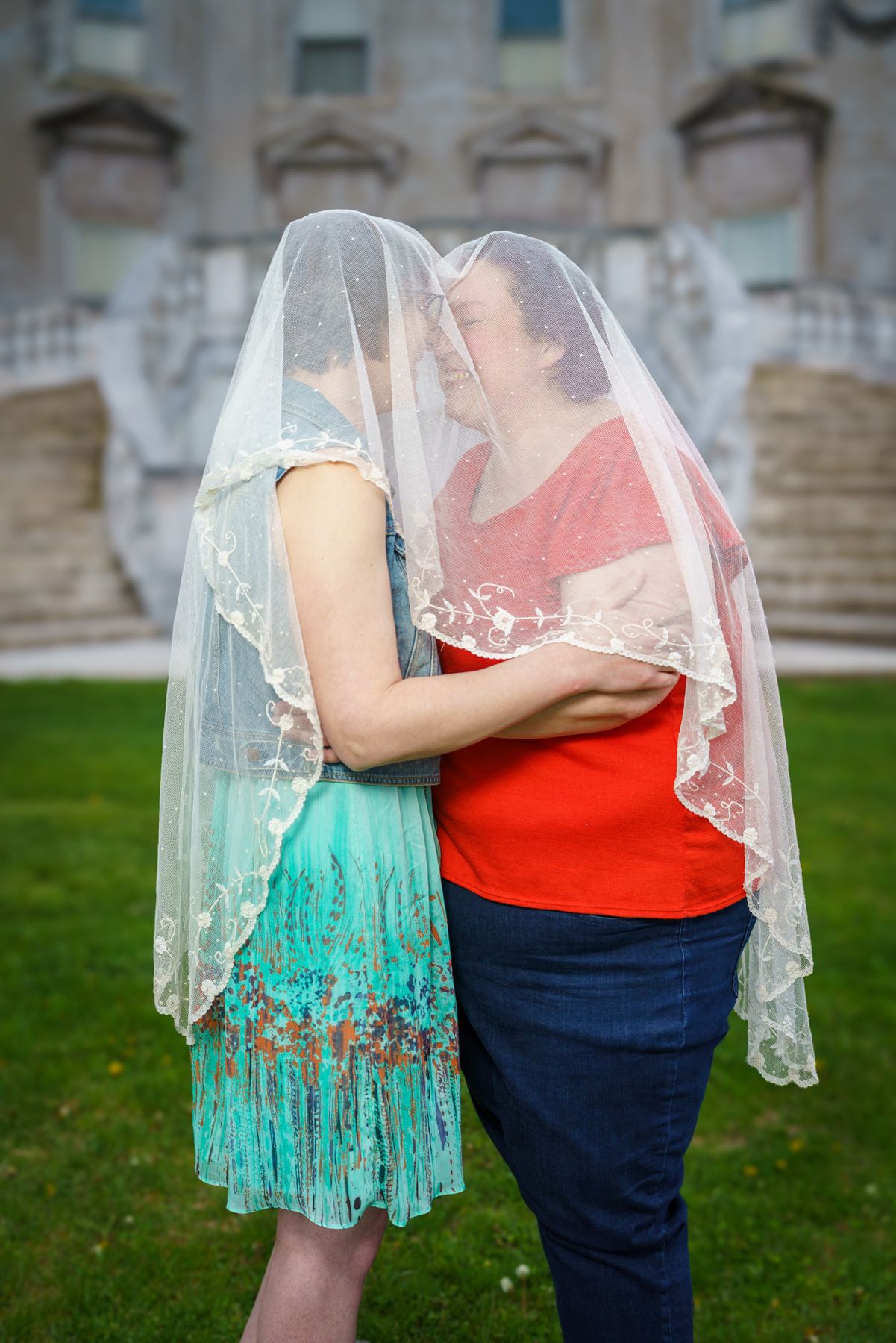 Two women share a romantic moment under a lace veil, embracing on a green lawn near stone steps.