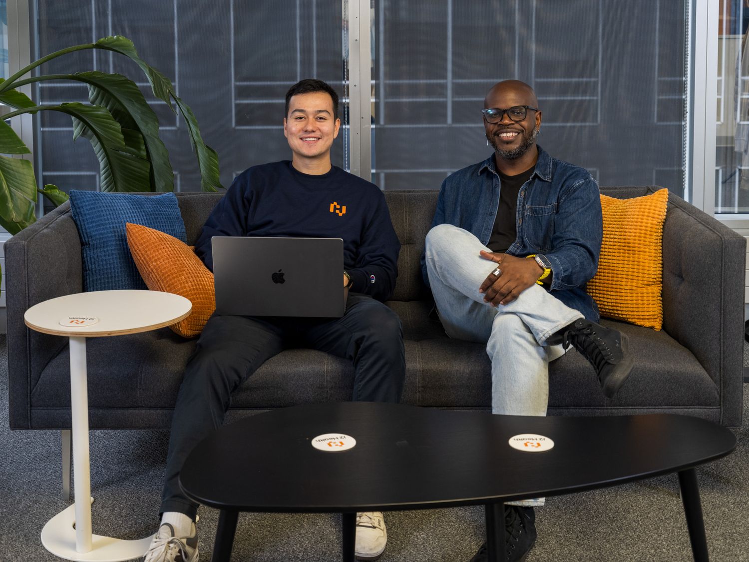 Two men sitting on a gray sofa with colorful pillows, one using a laptop, in a modern office lounge.
