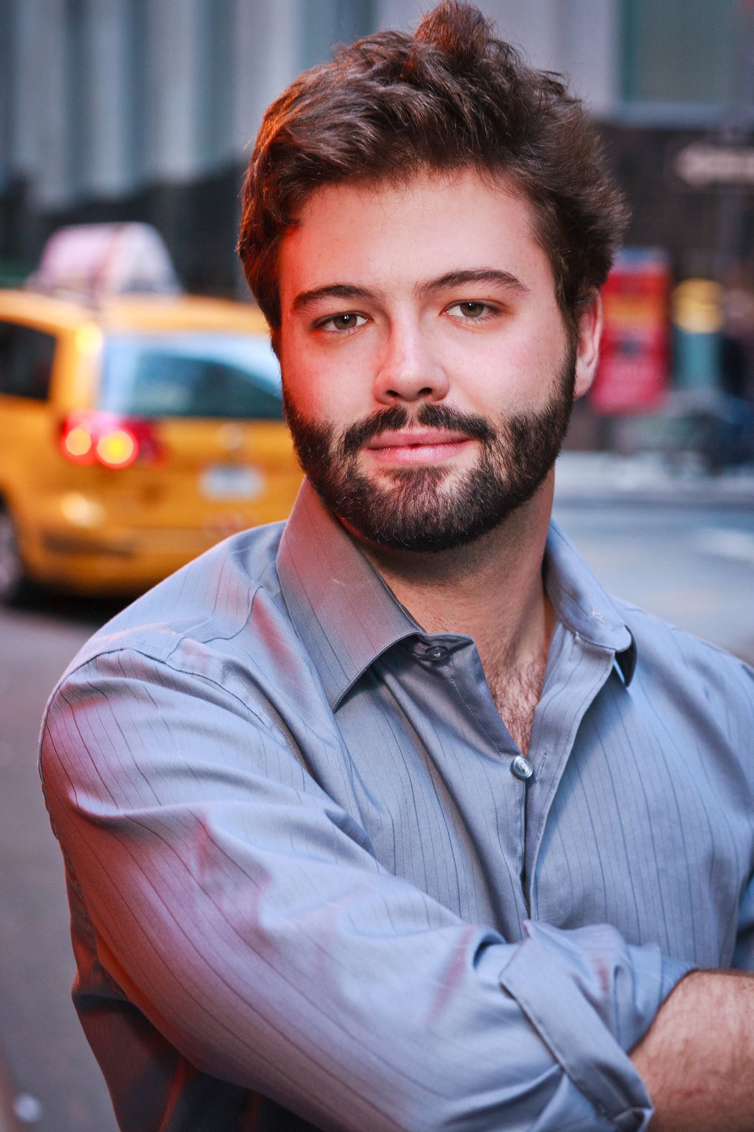Young bearded man with crossed arms posing outdoors on a city street with yellow taxi.