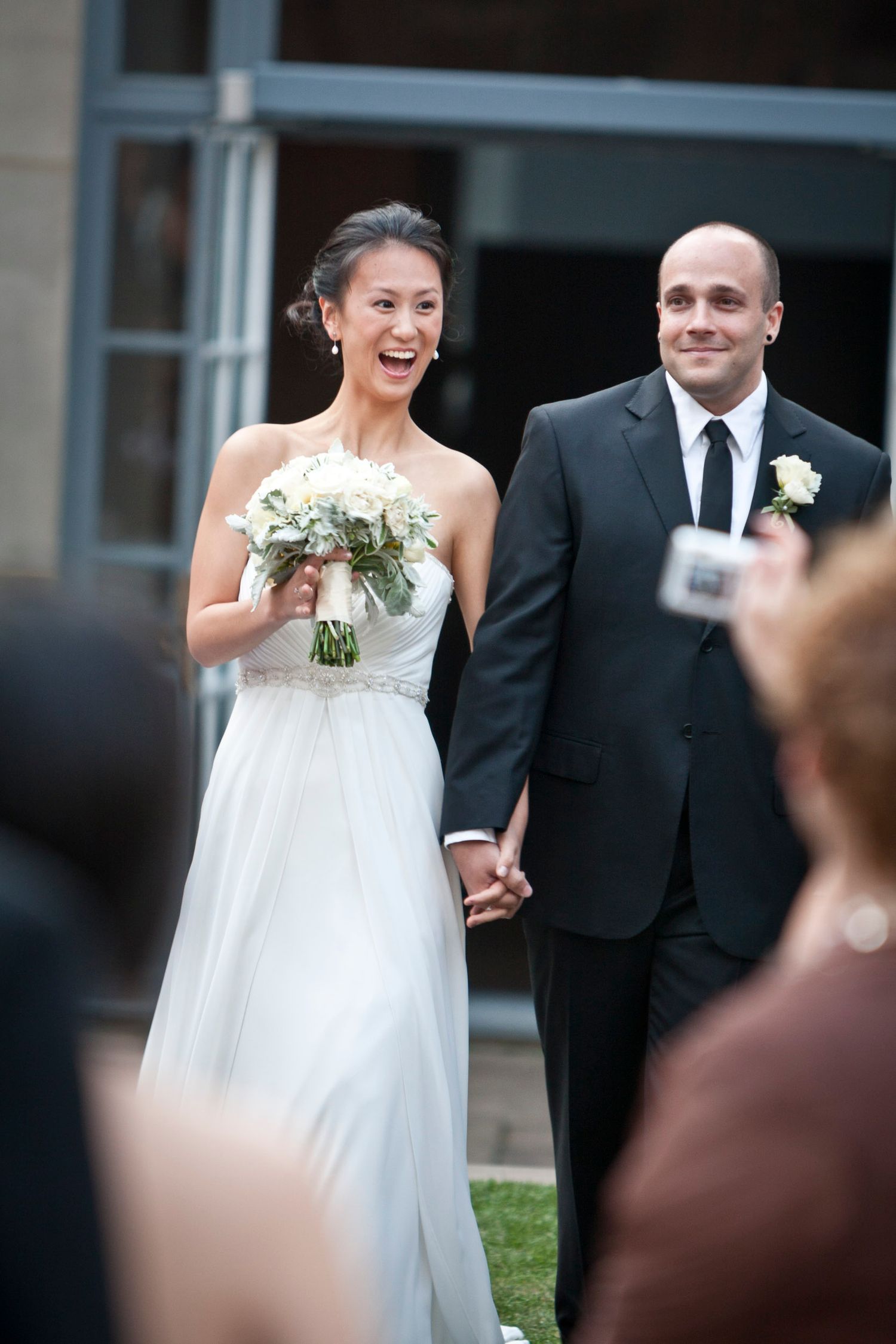 Smiling bride in white gown holding bouquet walks hand-in-hand with groom in black suit after wedding ceremony.