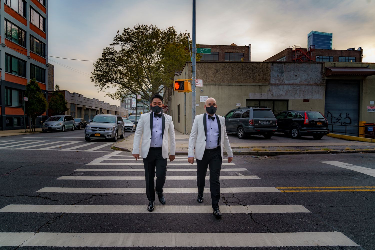 Two men in white tuxedos crossing a zebra crosswalk at sunset in the city.