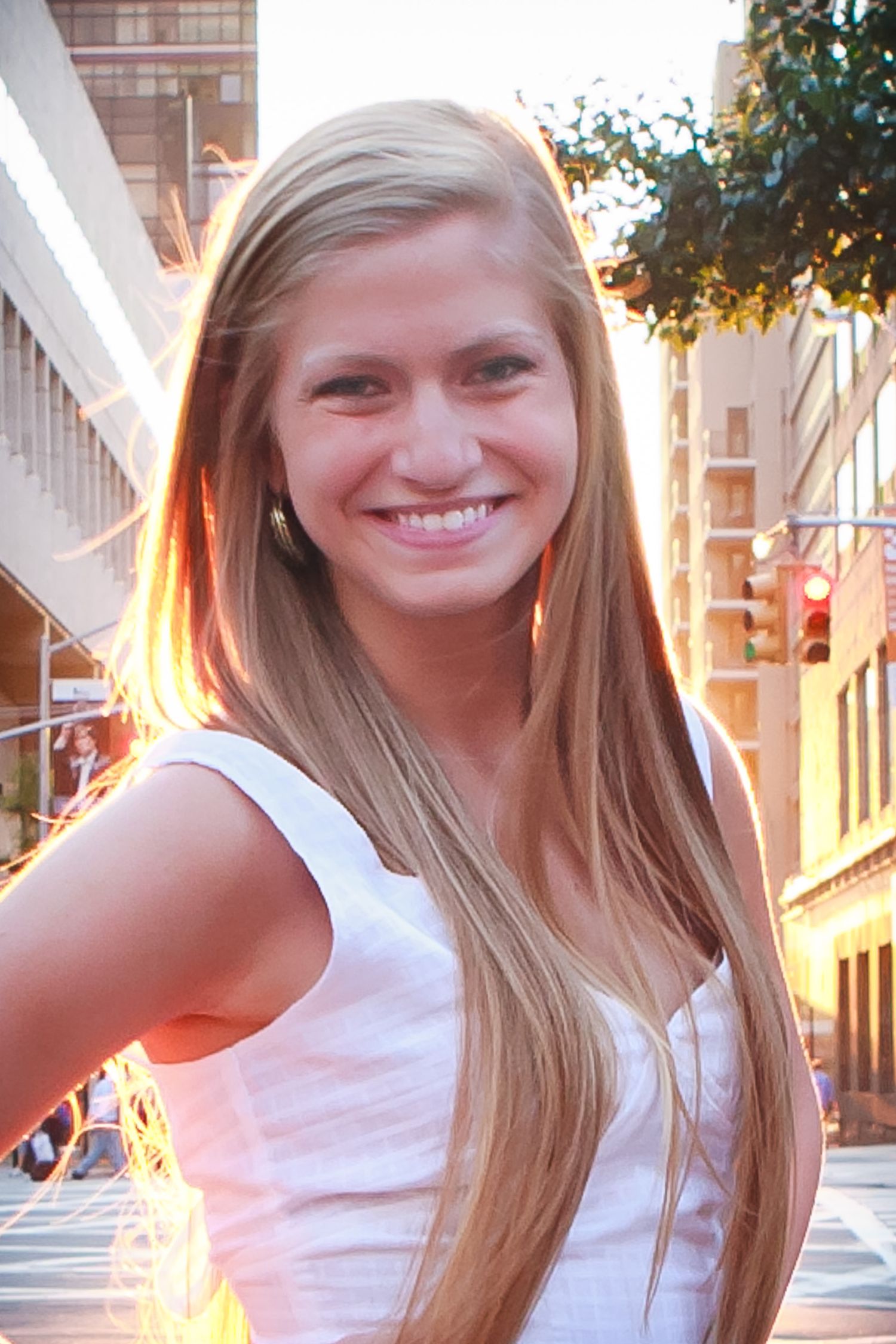 Smiling young blonde woman in white top posing on a sunny city street.