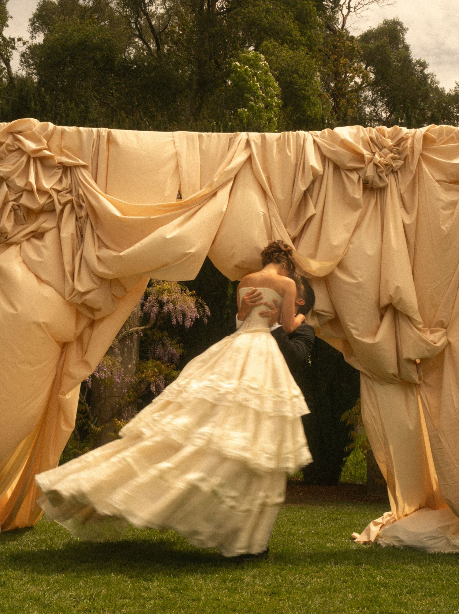 Bride in layered white gown kissing groom under elegant draped ivory fabric canopy at outdoor wedding ceremony.