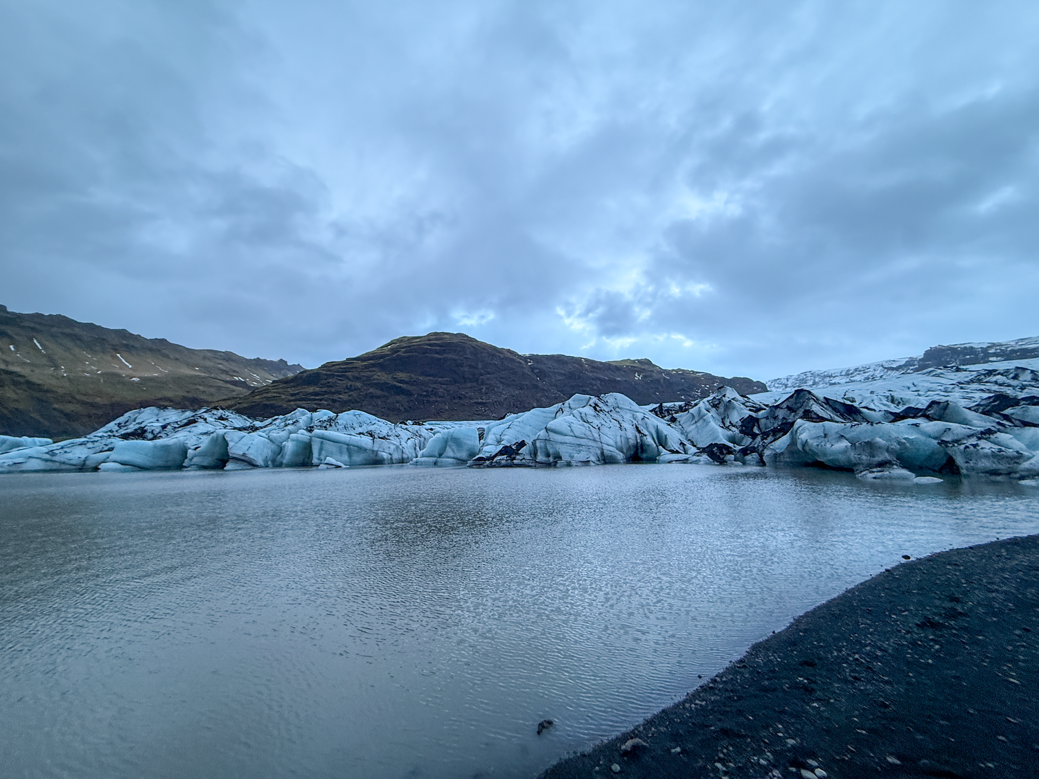 Glacial lagoon with floating icebergs, black volcanic shoreline, and dramatic cloudy skies in Iceland.