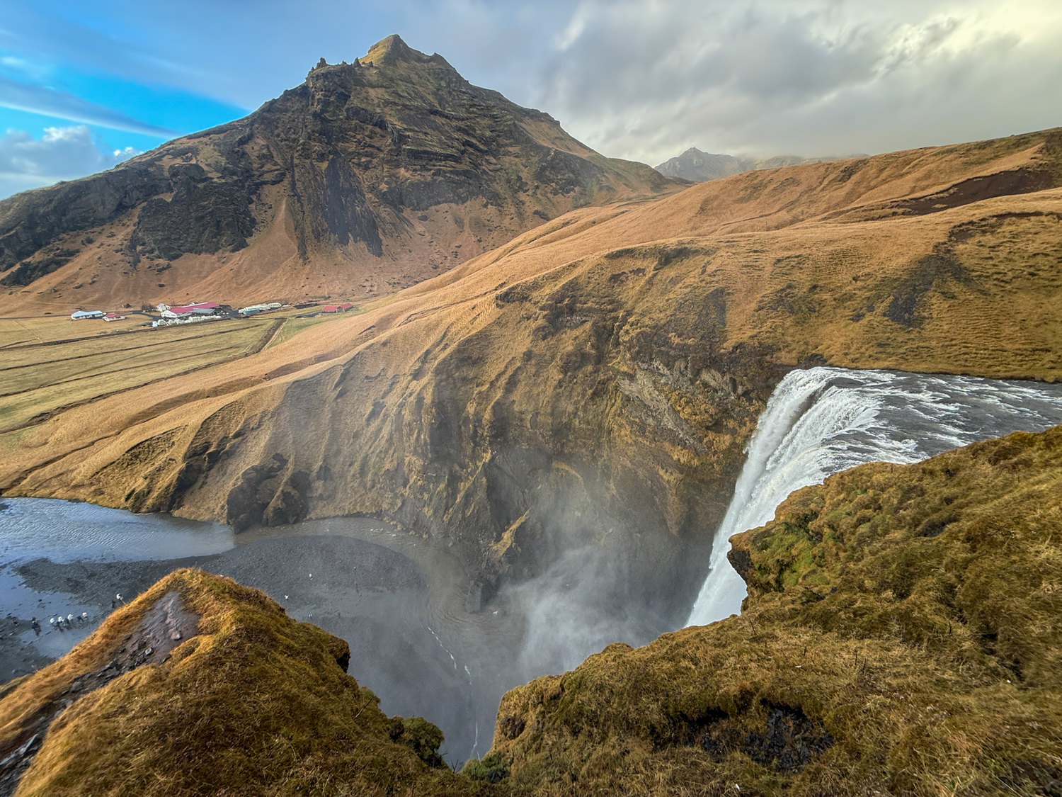 Aerial view of a waterfall in Iceland with misty spray, golden hillsides, and a dramatic mountain peak backdrop.