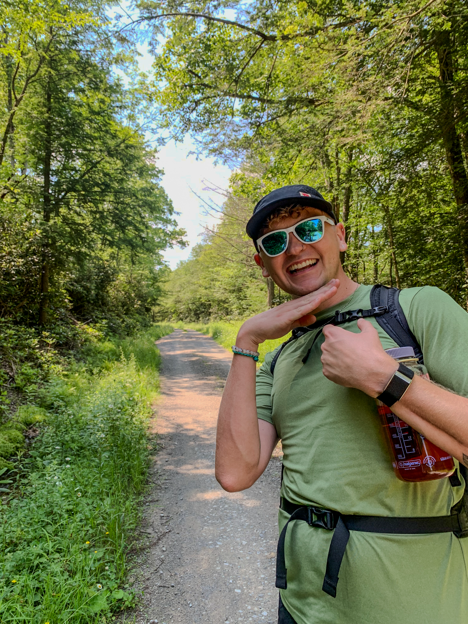 Smiling hiker with sunglasses and green shirt poses on a lush forest trail while carrying a backpack.