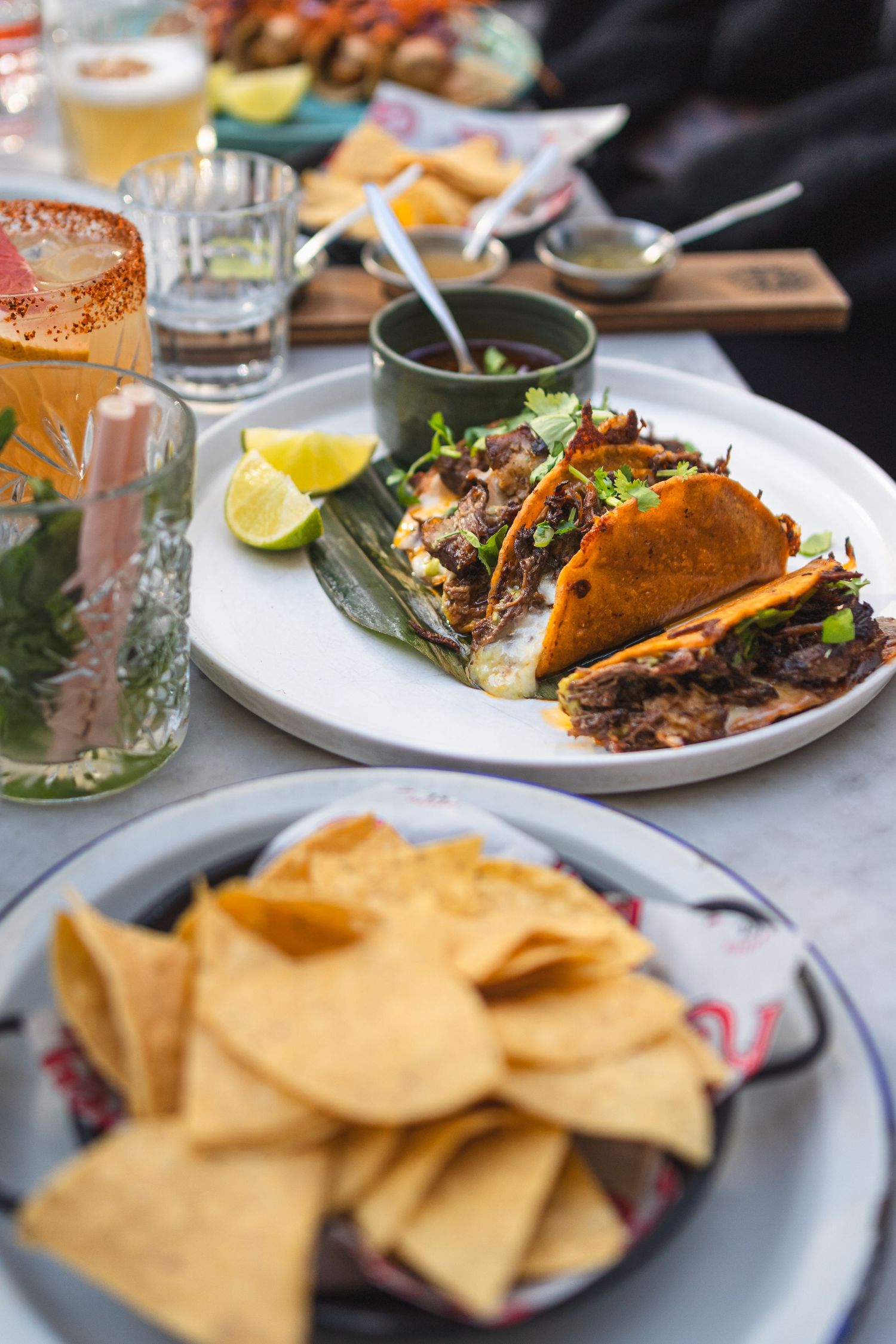 Mexican tacos with pulled meat and chips with lime wedges and drinks on restaurant table.