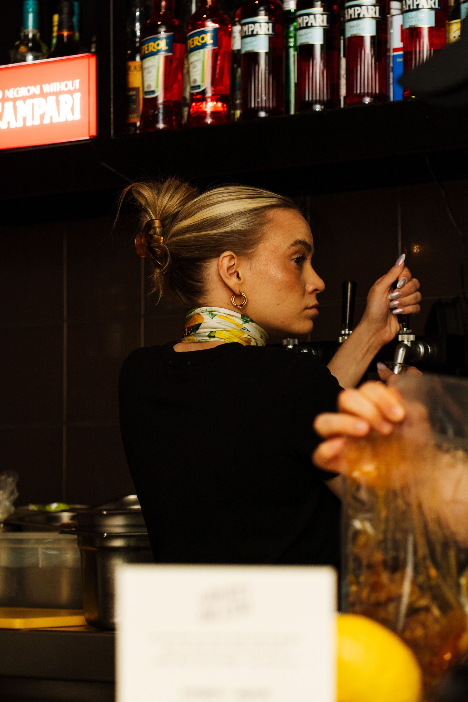 Female bartender pulling draft beer tap at dimly lit bar with Campari bottles.