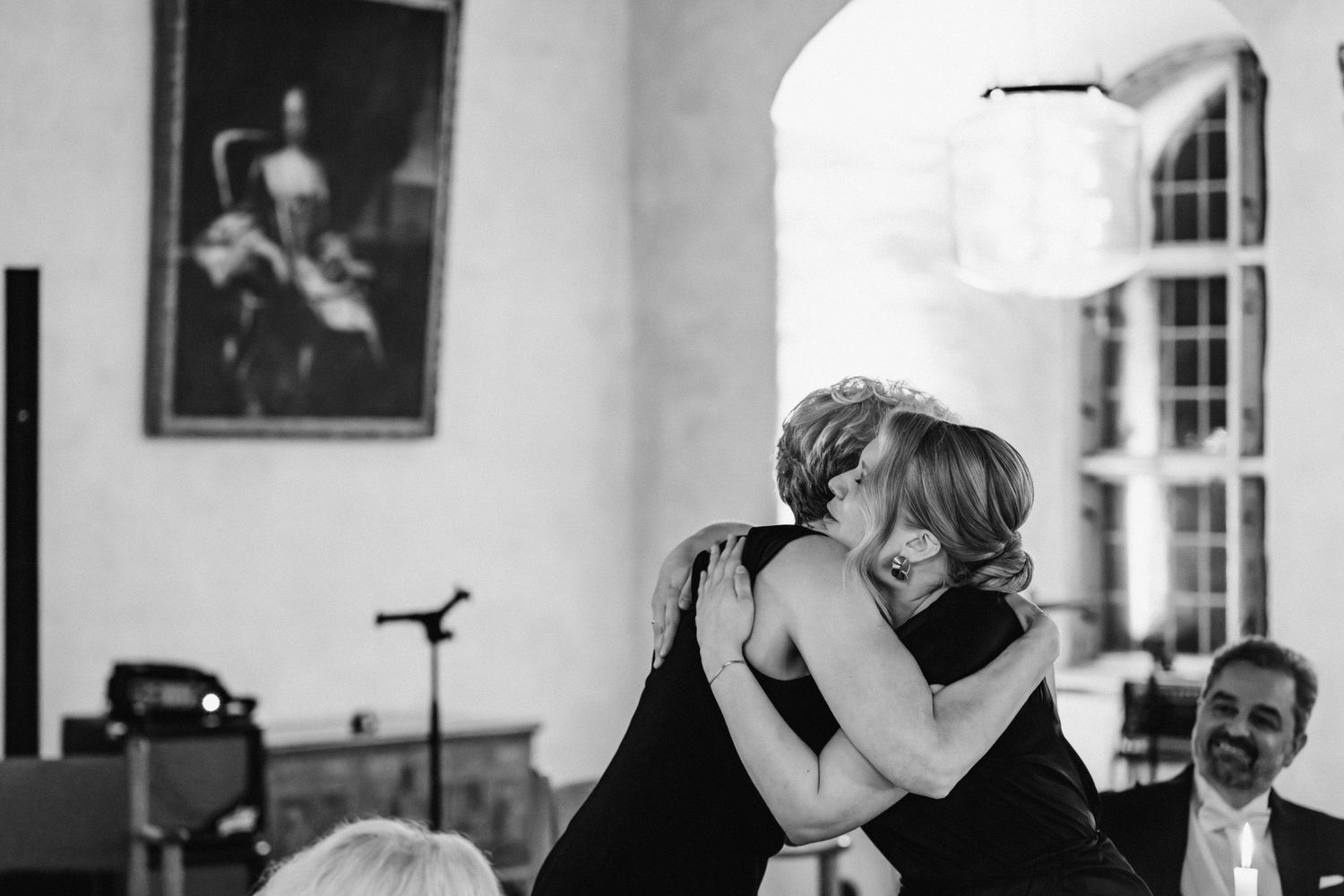 Two women share an emotional hug at a formal event with a portrait painting on the wall behind them.