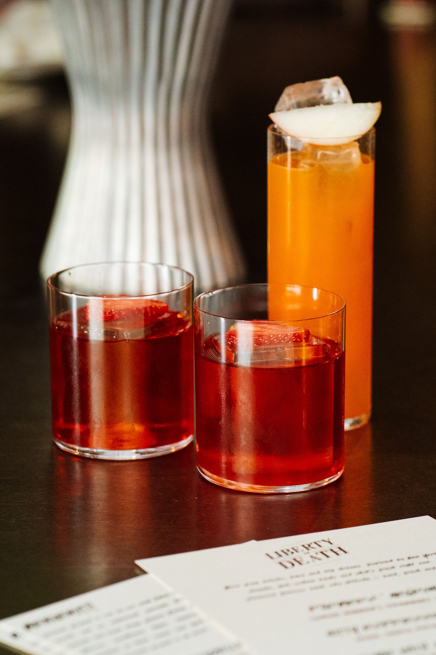 Two red Negroni cocktails and an orange drink on a wooden bar next to a Liberty Death menu.