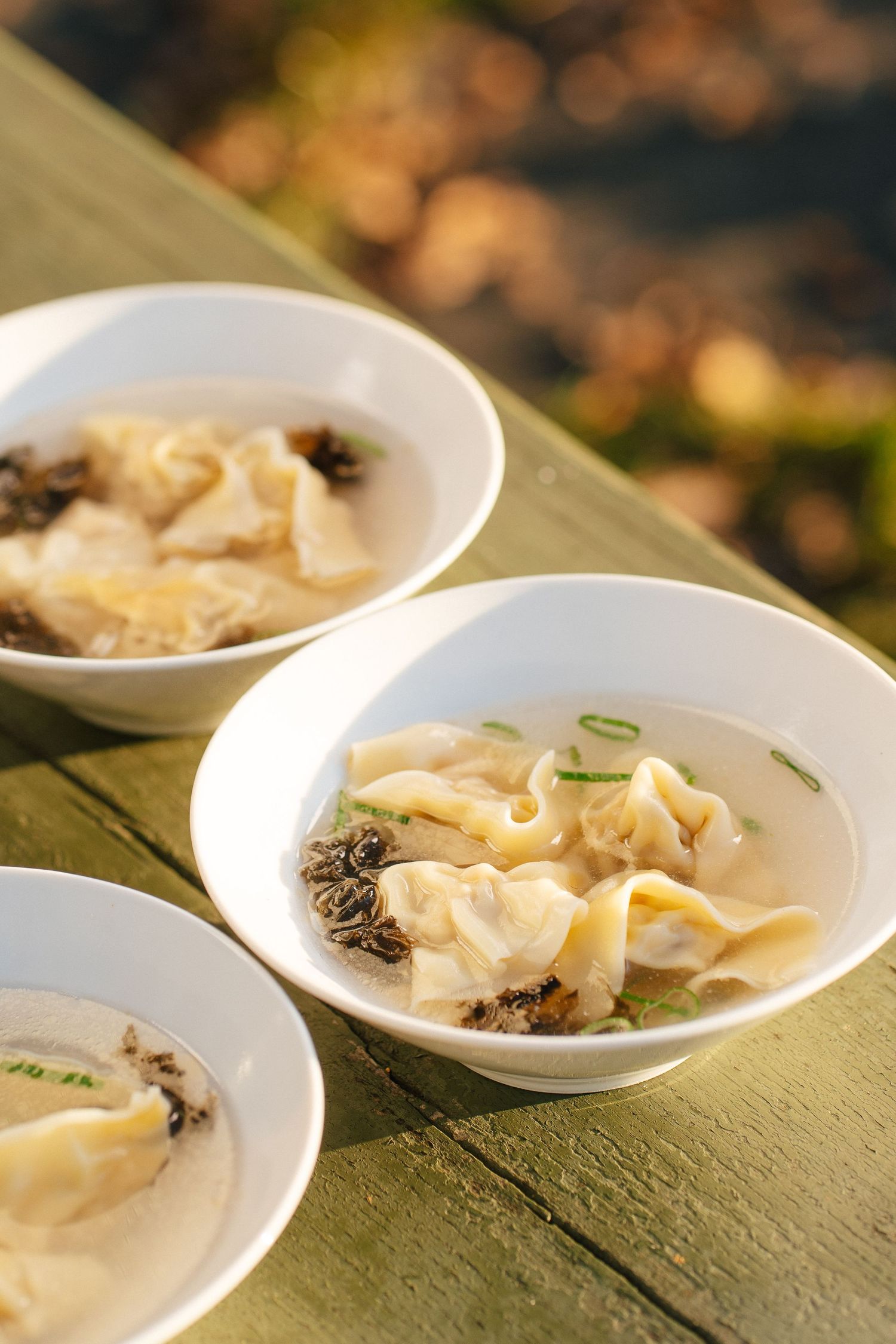 Three white bowls of wonton soup with mushrooms served on green wooden surface.
