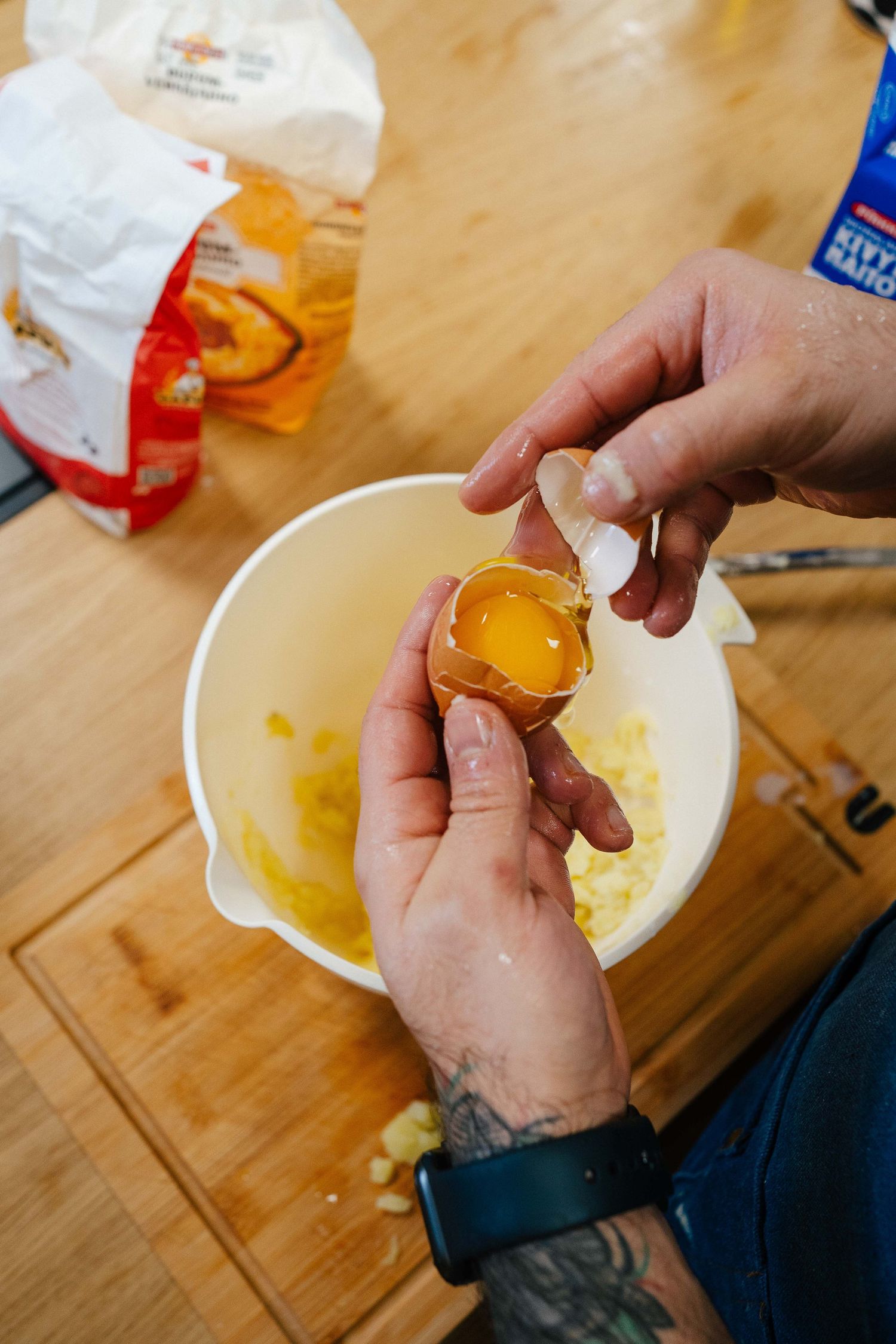 Tattooed hands cracking a fresh egg into a white mixing bowl with mashed potatoes on a wooden board.