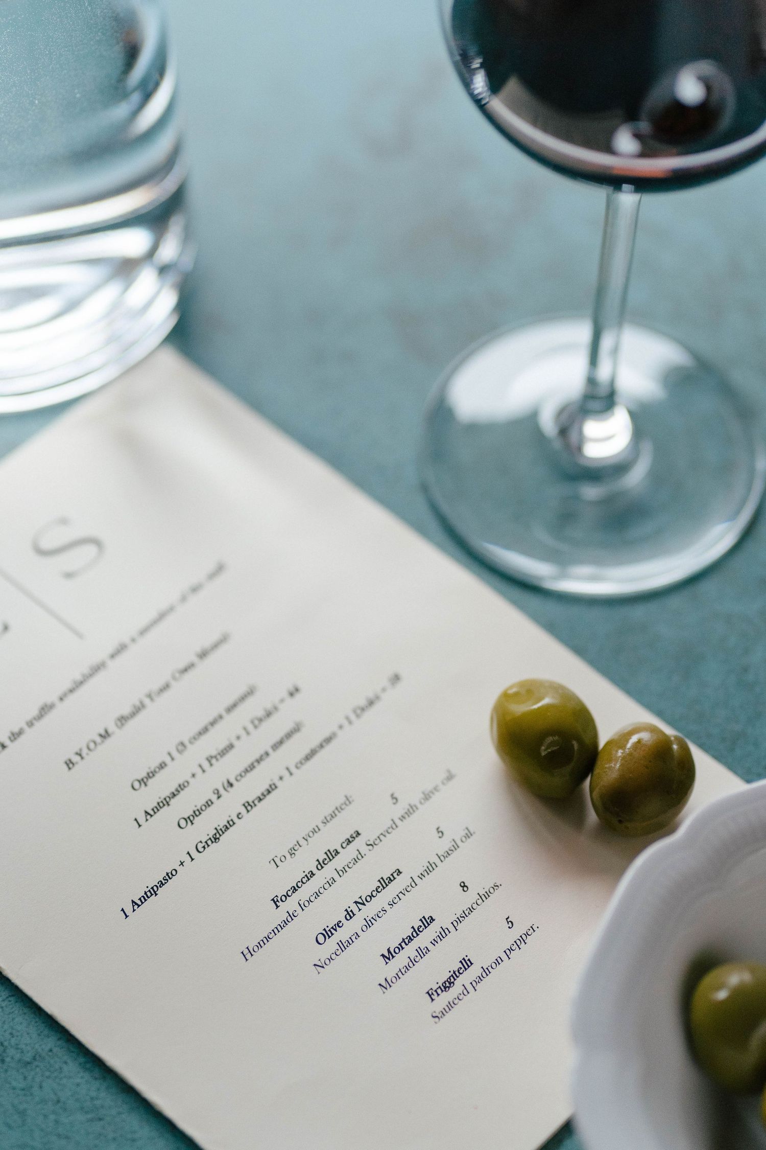 Restaurant menu with green olives and glass of red wine on teal table surface.