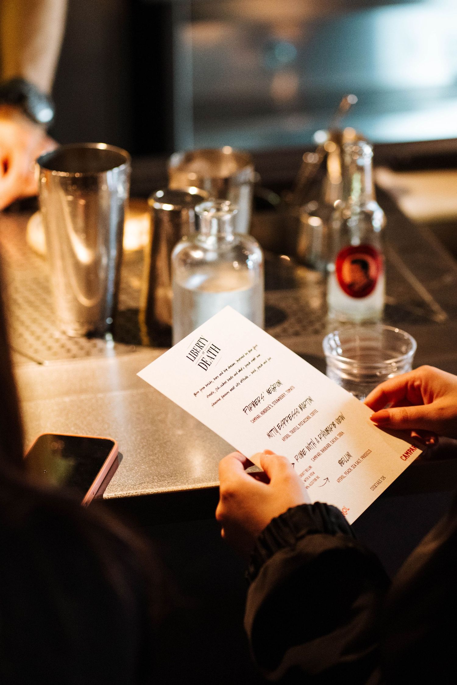 Person holding a cocktail menu at a bar with shakers and glass bottles in the background.