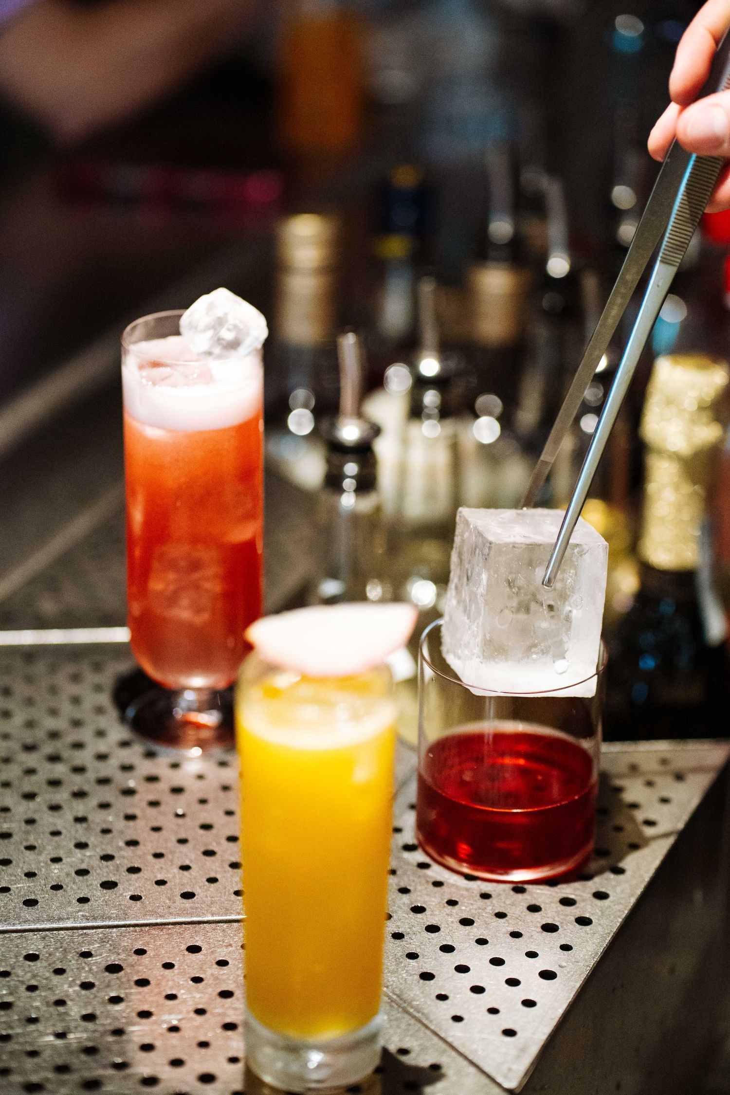 Bartender using tongs to place a large ice cube into a red cocktail glass at a bar.