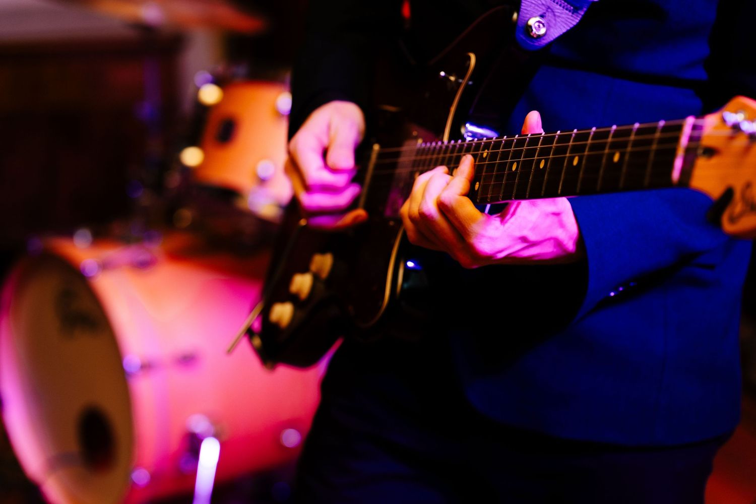 Close-up of musician playing electric guitar on stage with colorful pink and purple lighting.