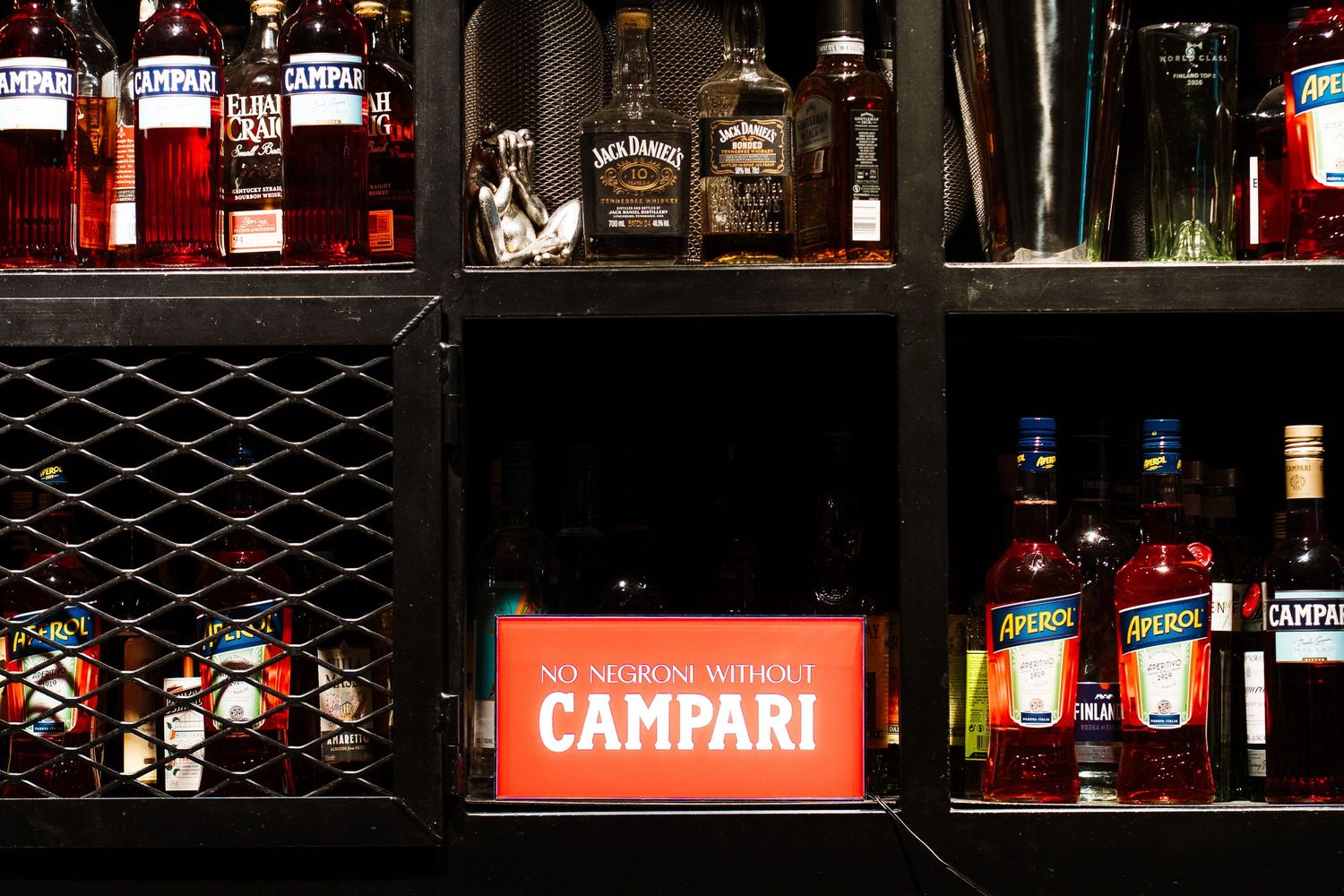 Bar shelf displaying Campari bottles with a glowing sign reading No Negroni Without Campari.