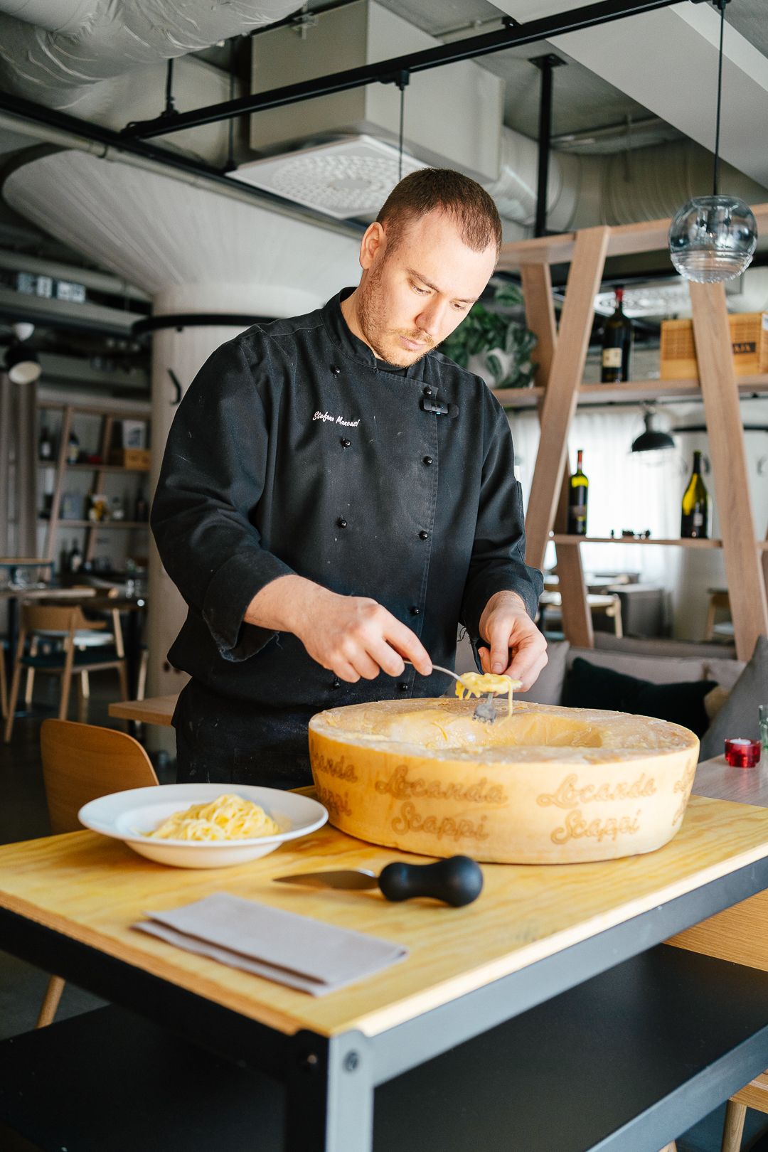 Chef in black uniform preparing pasta inside a hollowed Parmigiano Reggiano wheel at a restaurant.